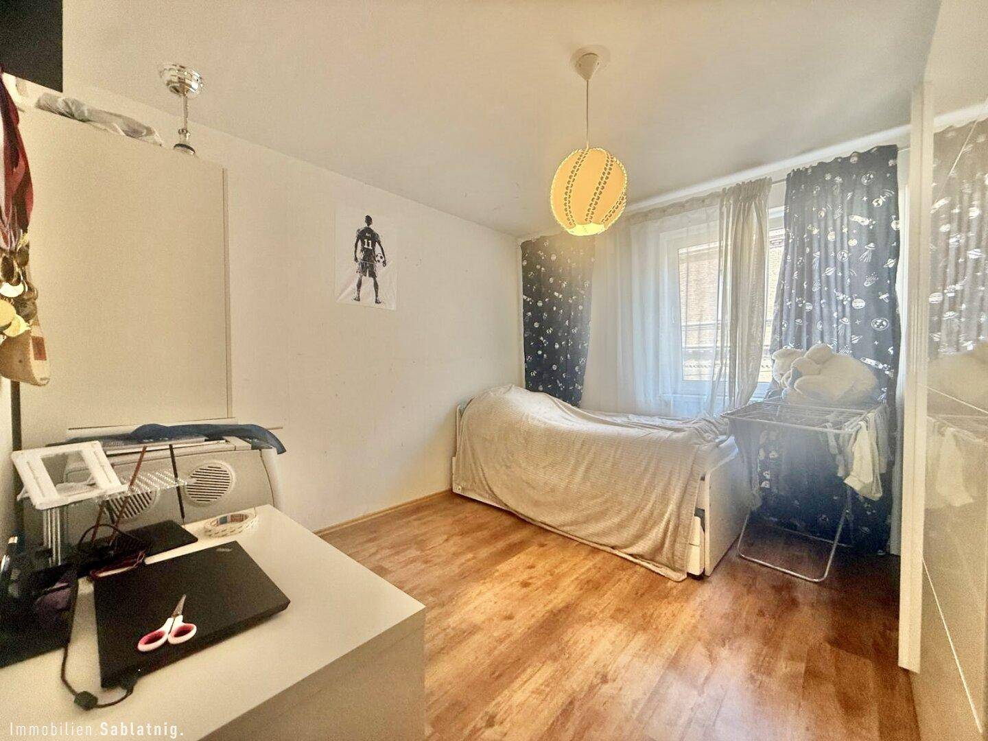 Einfaches Schlafzimmer mit Holzboden, Fenster und funktionaler Einrichtung.
