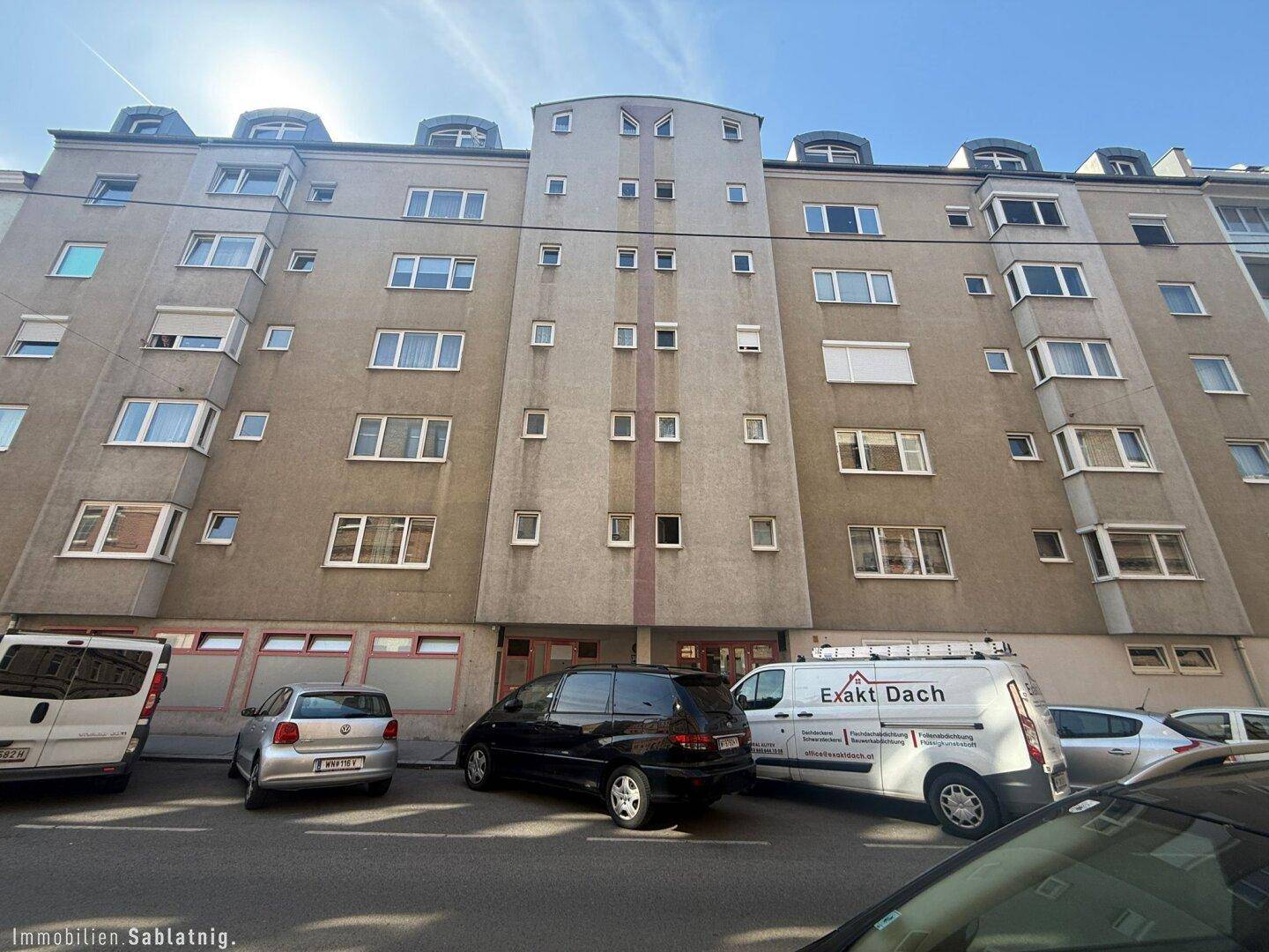 Mehrfamilienhaus mit moderner Fassade, vielen Fenstern und Parkplätzen an der Straße.