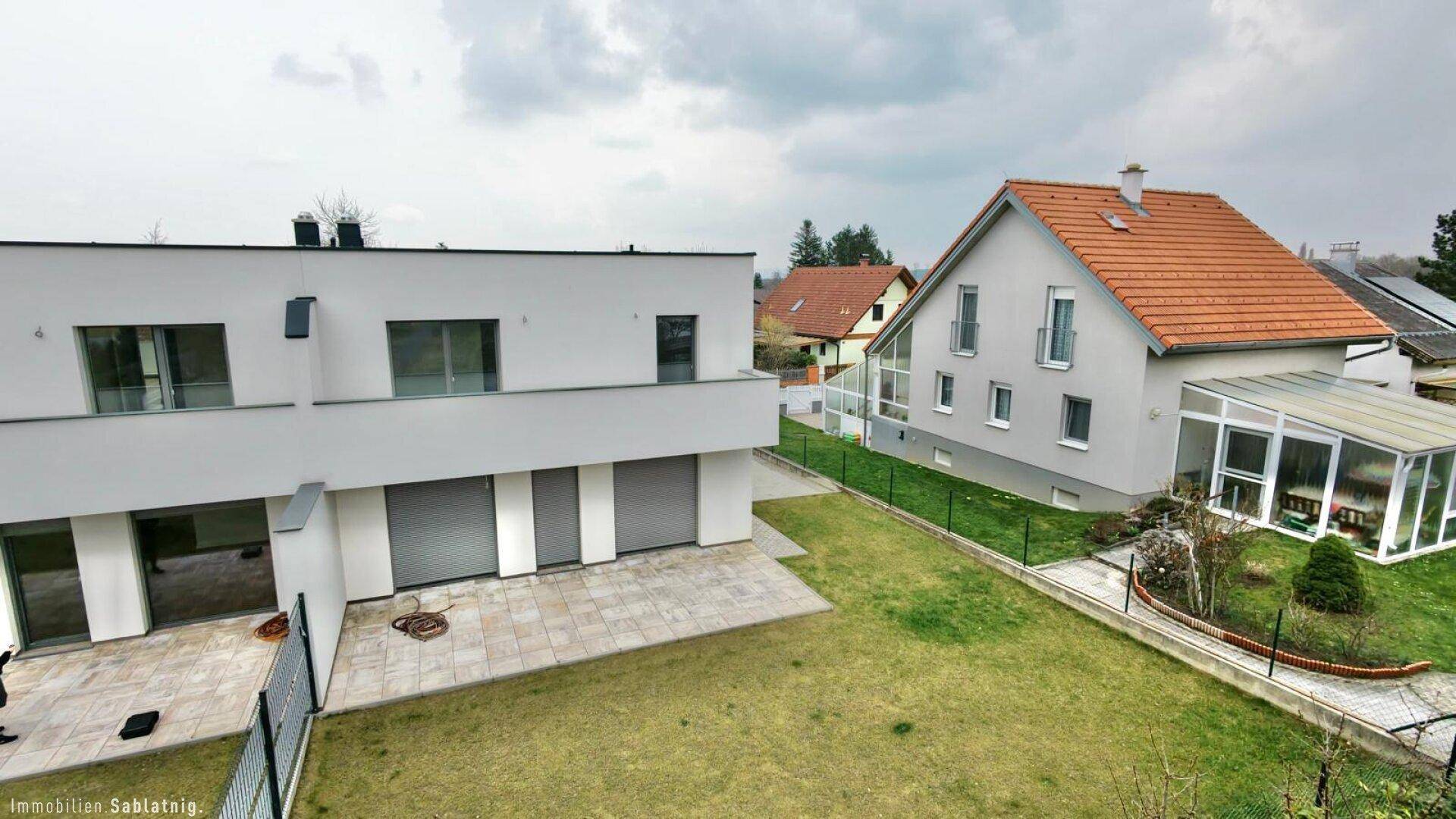 Rückansicht des Neubaus mit Terrasse und Garten, angrenzend an ein älteres Haus mit Wintergarten.