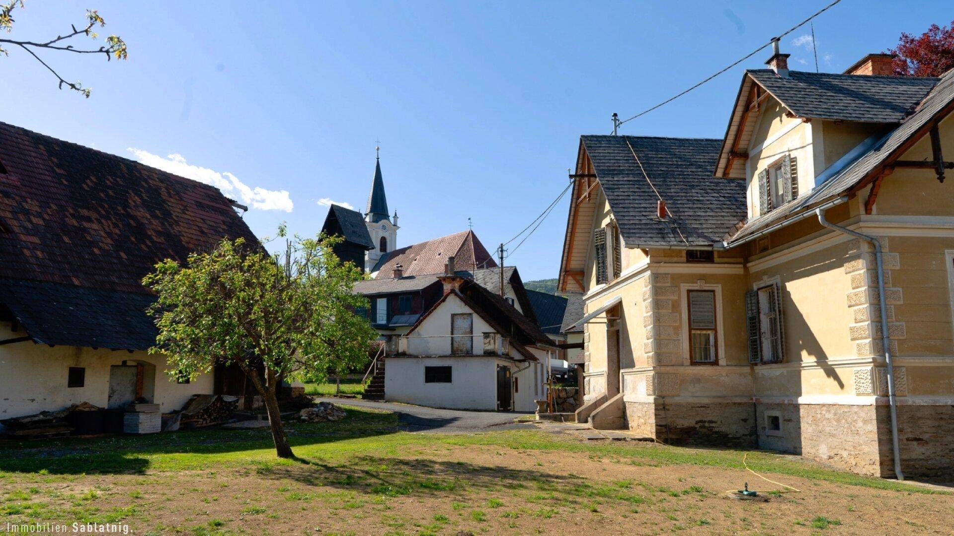Anwesen mit mehreren Gebäuden und der Dorfkirche im Hintergrund, umgeben von Grünflächen.