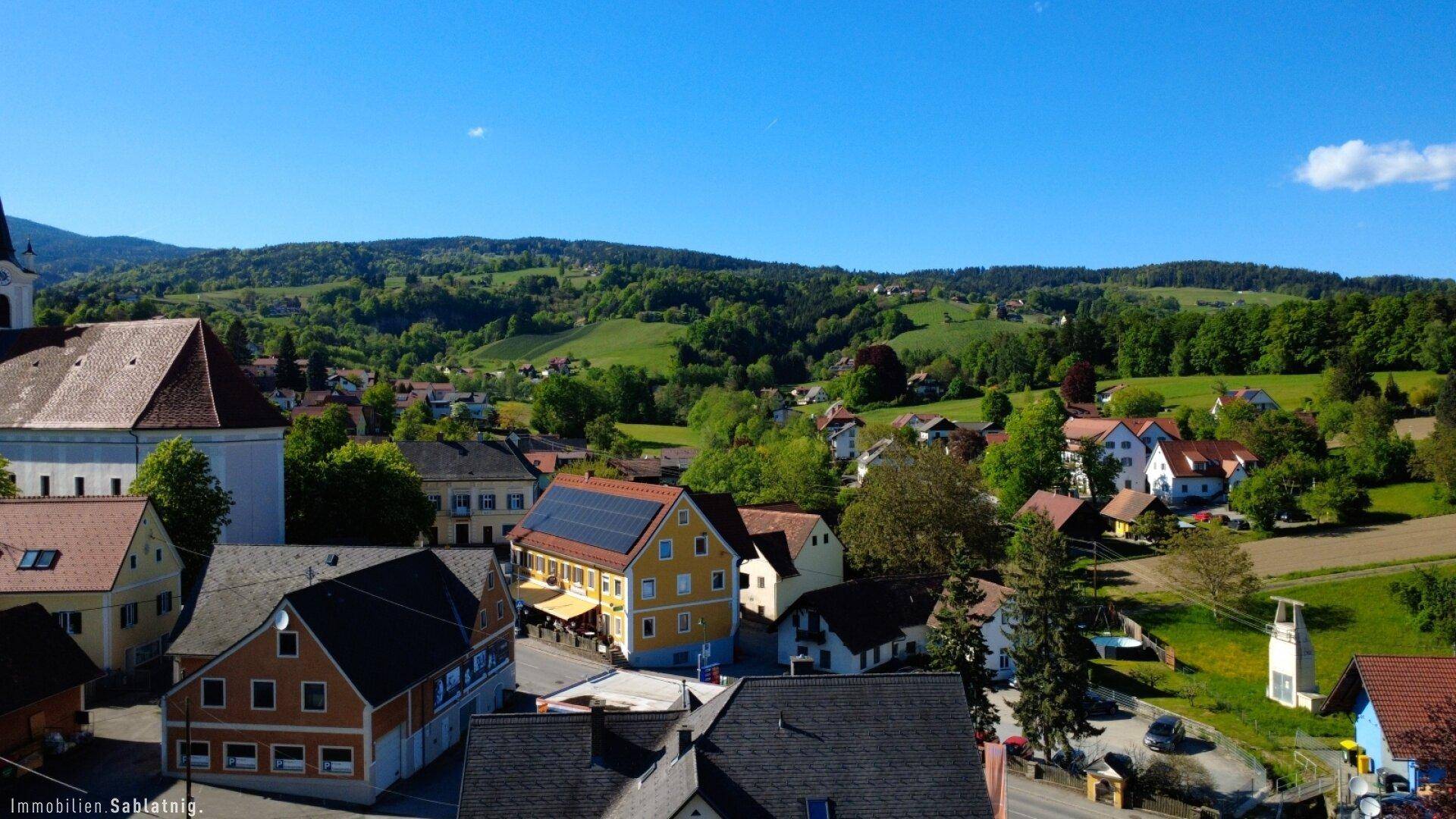 Panoramablick auf das Dorf mit grünen Hügeln und Feldern unter einem weiten blauen Himmel.