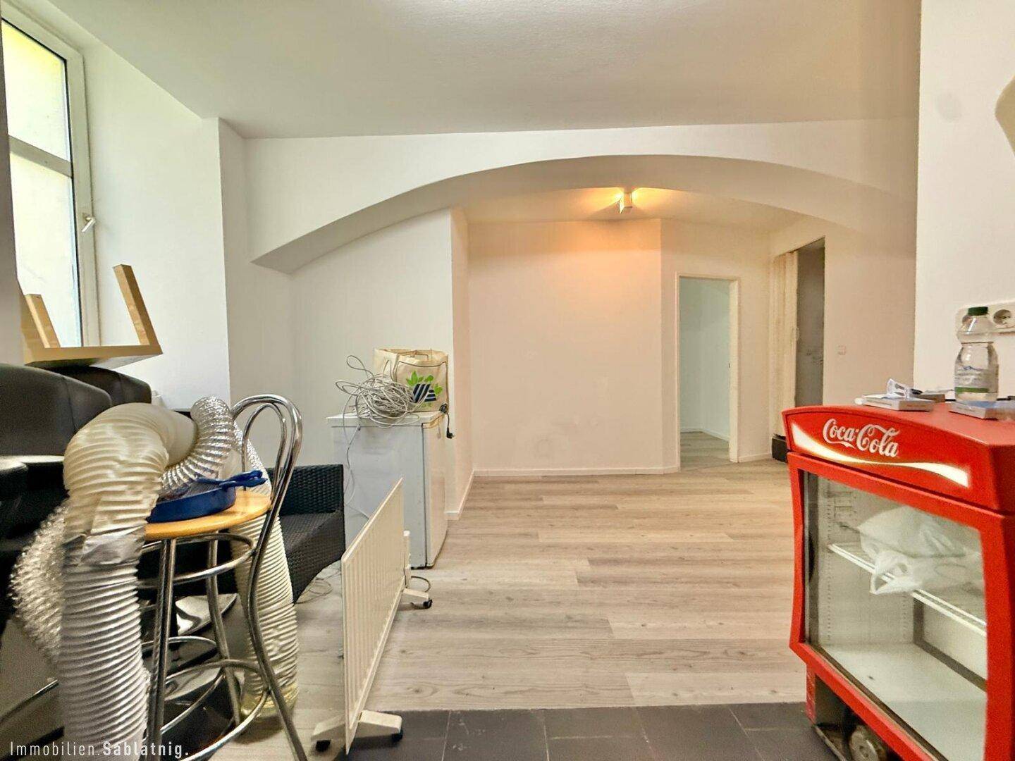 Offener Wohnbereich mit hellem Boden und gewölbter Decke, im Hintergrund ein Fenster und ein Coca-Cola Kühlschrank.