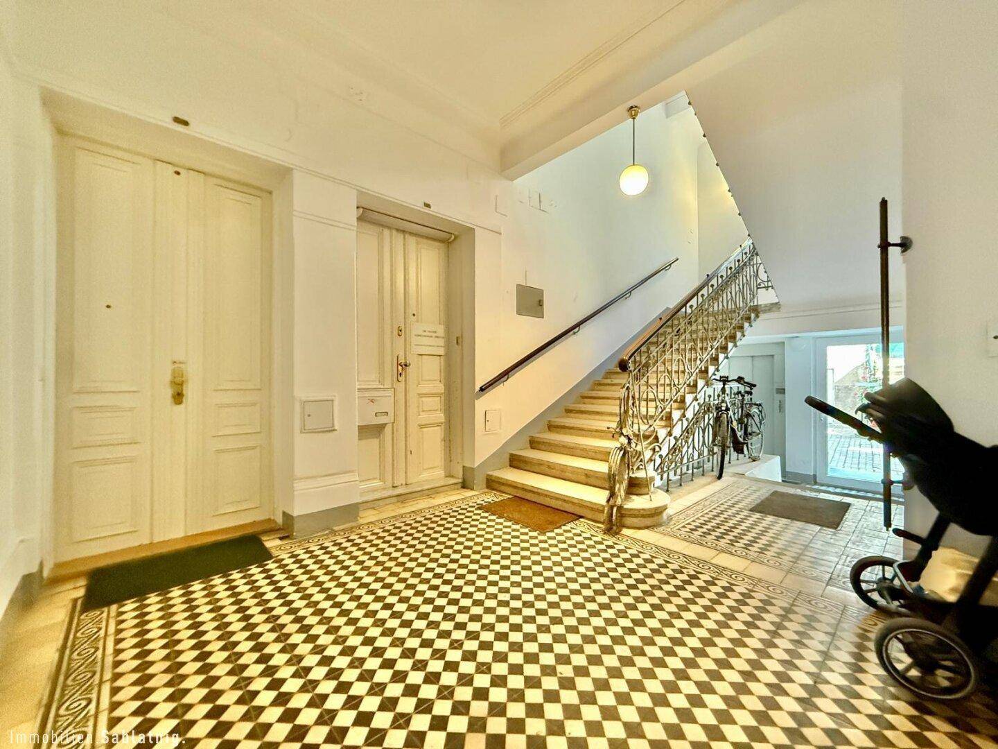 Historischer Eingangsbereich mit Schachbrettmuster-Fliesen und einer eleganten Treppe mit schmiedeeisernem Geländer.