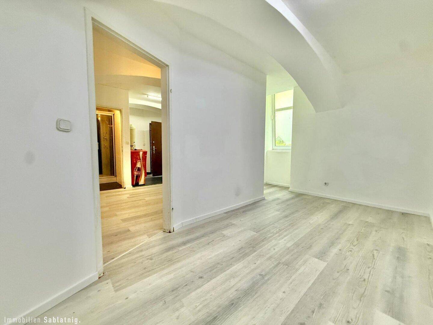 Heller Wohnbereich mit Laminatboden und gewölbter Decke, ein Durchgang führt zu einem weiteren Raum.