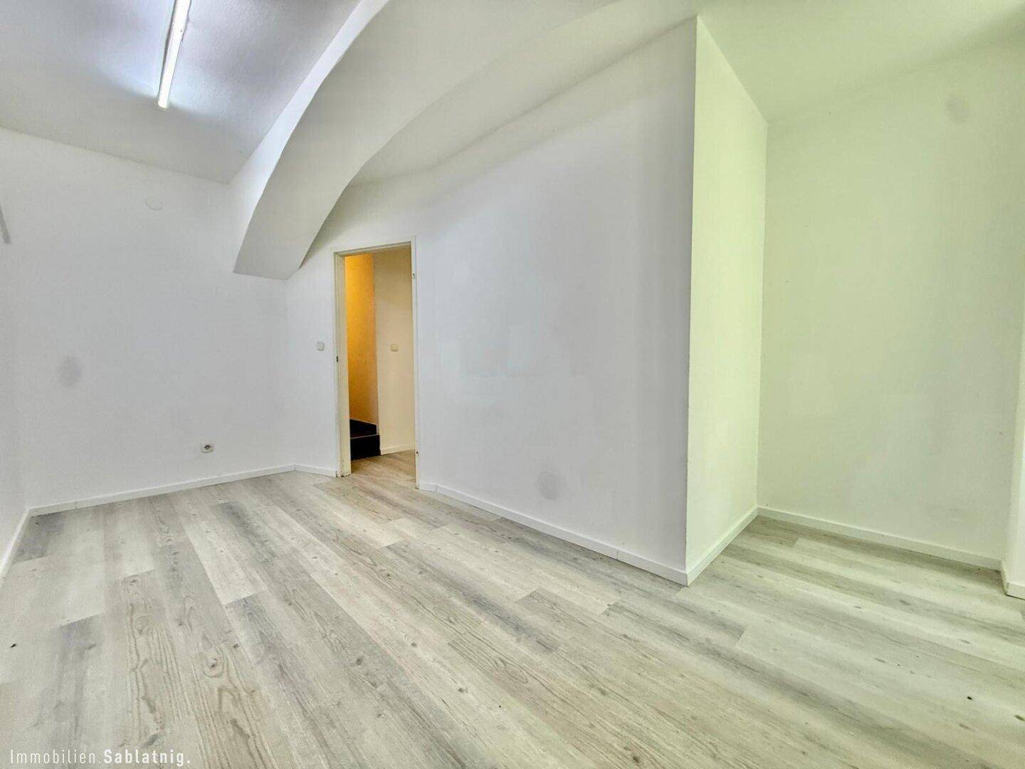 Heller Raum mit Laminatboden und weiß gestrichenen Wänden, ein Durchgang führt in einen angrenzenden Bereich.
