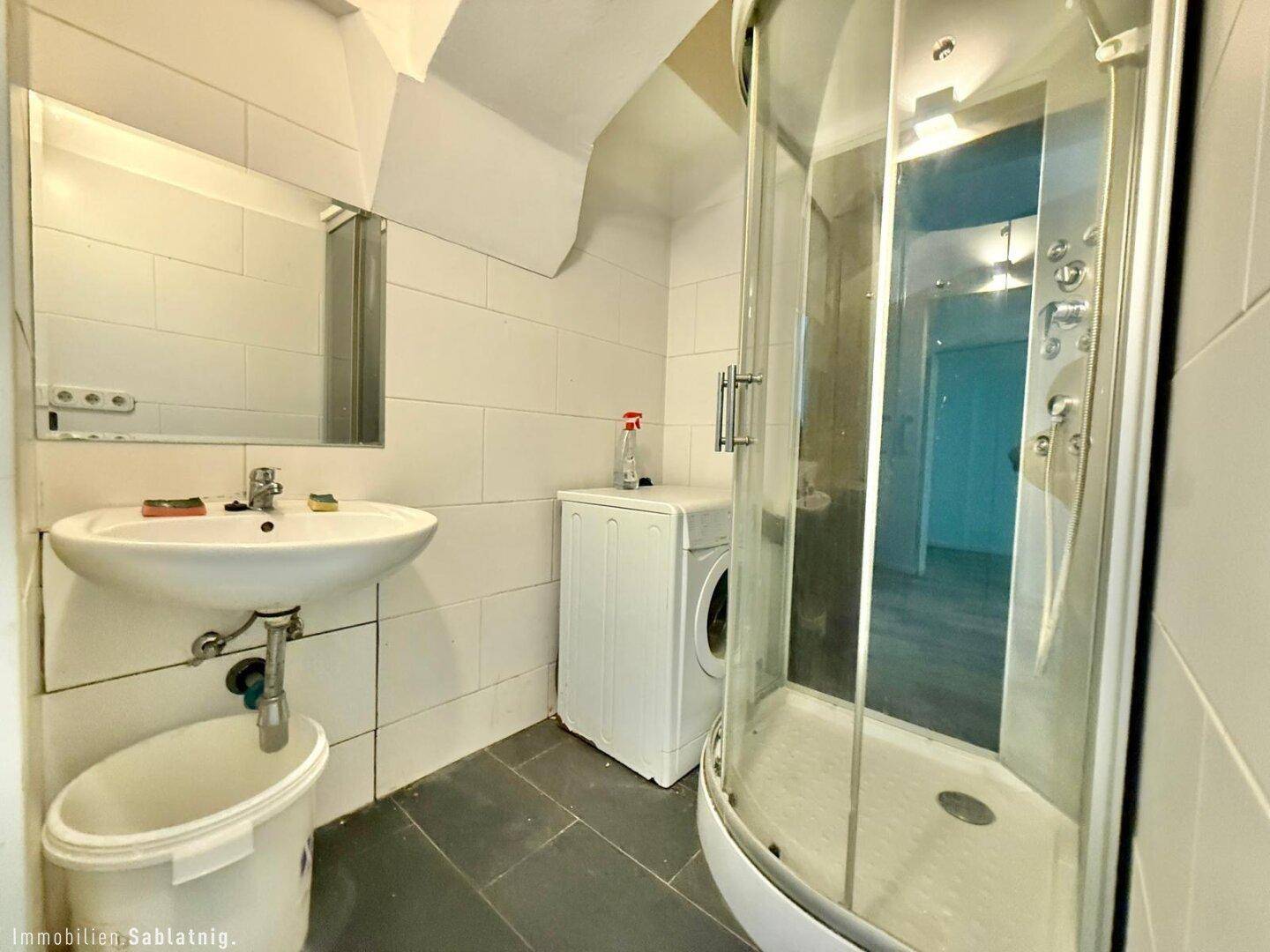 Badezimmer mit Duschkabine, Waschbecken und Waschmaschine, die Wände sind weiß gefliest.
