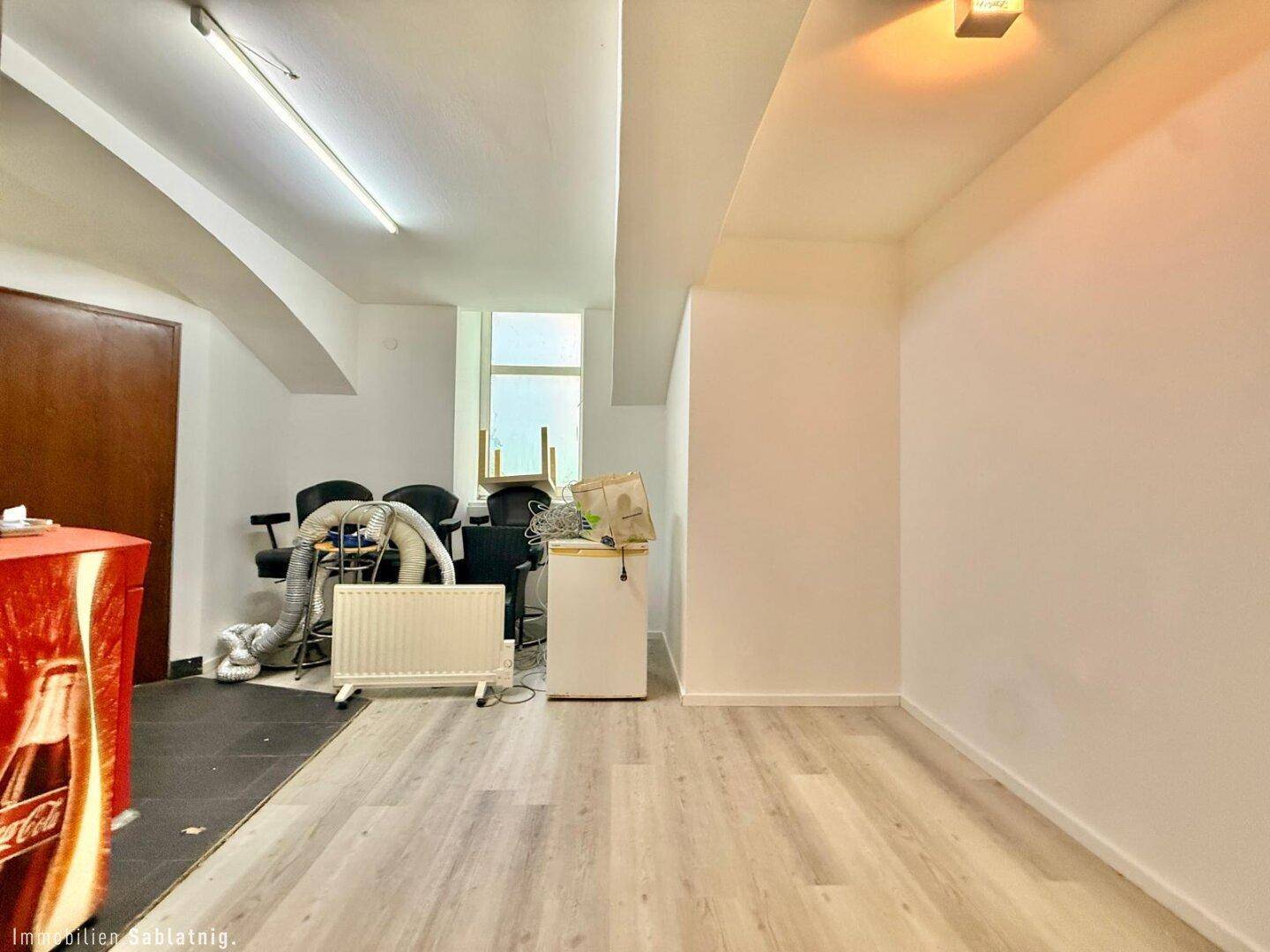 Heller Wohnraum mit Laminatboden und einem Fenster, im Hintergrund sind Stühle und ein Kühlschrank zu sehen.