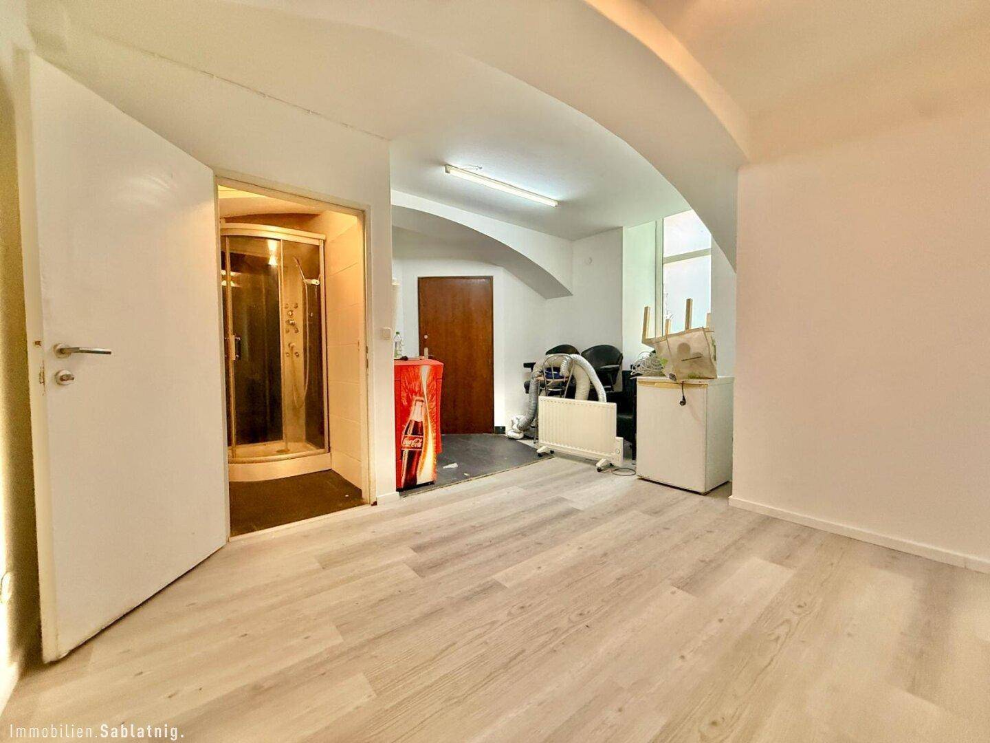 Geräumiger Wohnbereich mit hellem Laminatboden, Blick auf eine Duschkabine und einen roten Kühlschrank.