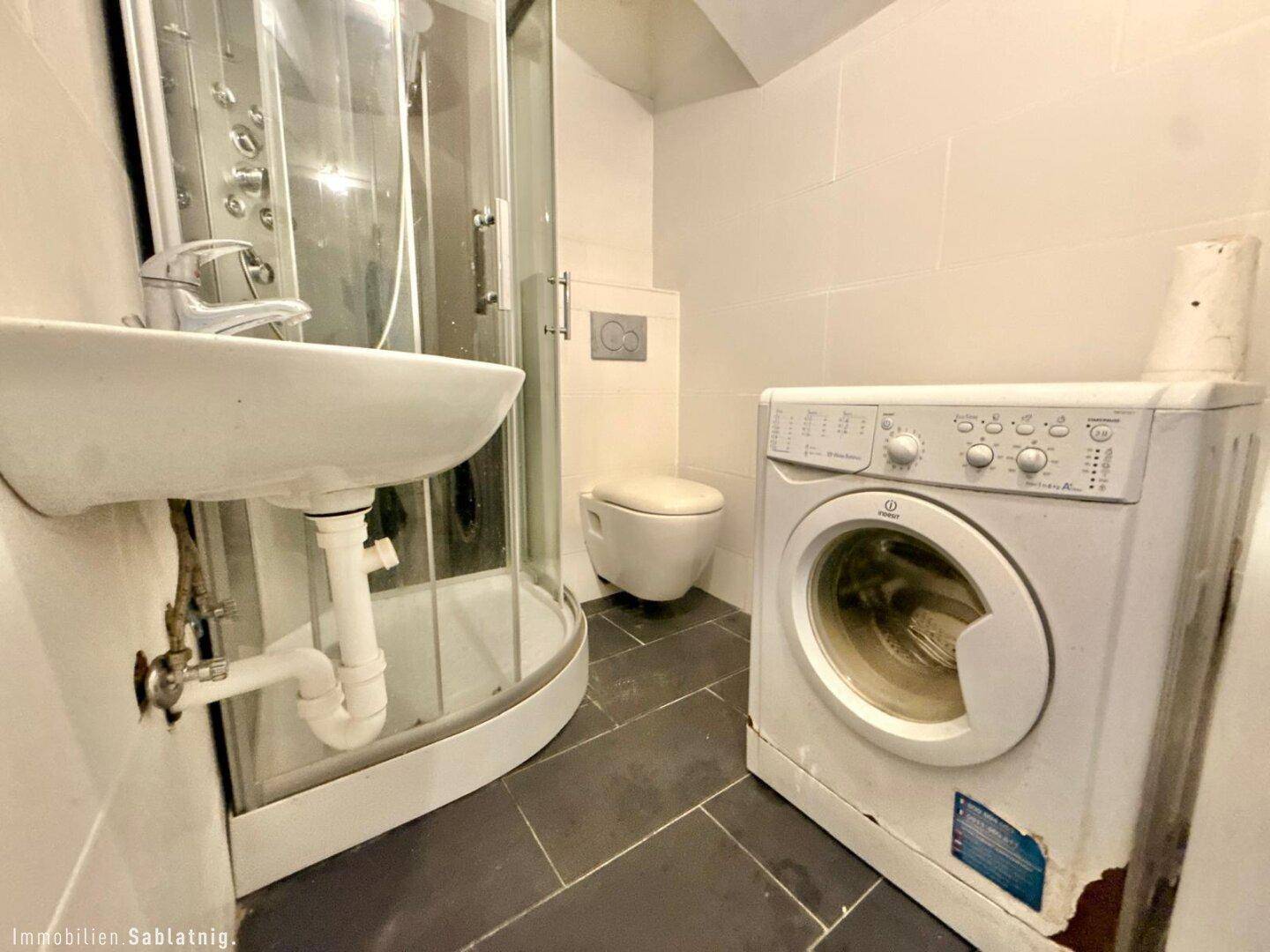 Badezimmer mit Duschkabine, Waschbecken und Waschmaschine, im Hintergrund ist eine Toilette sichtbar.
