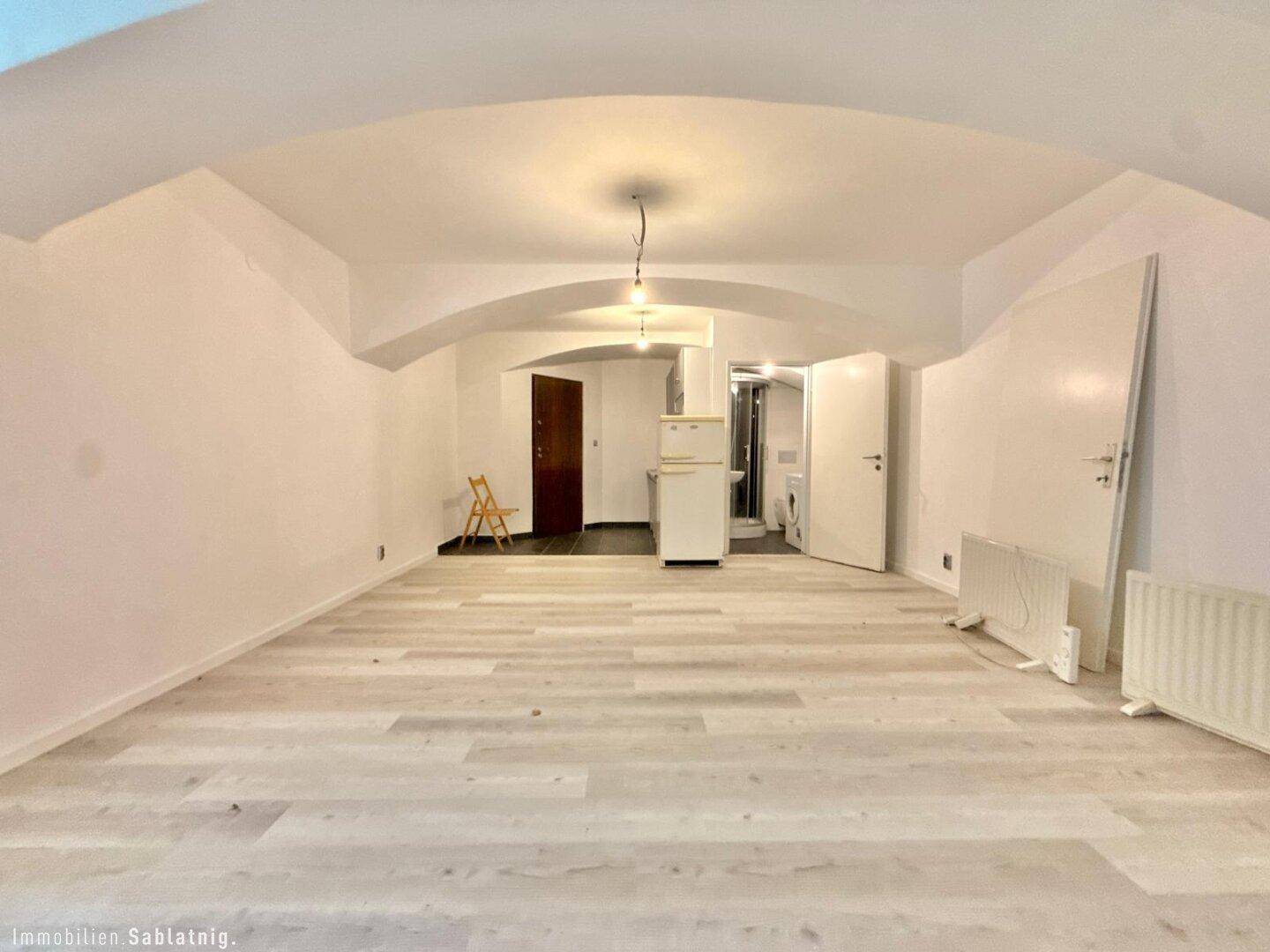 Heller Wohnraum mit Laminatboden und gewölbter Decke, Blick auf den Eingangsbereich und eine kleine Küche.
