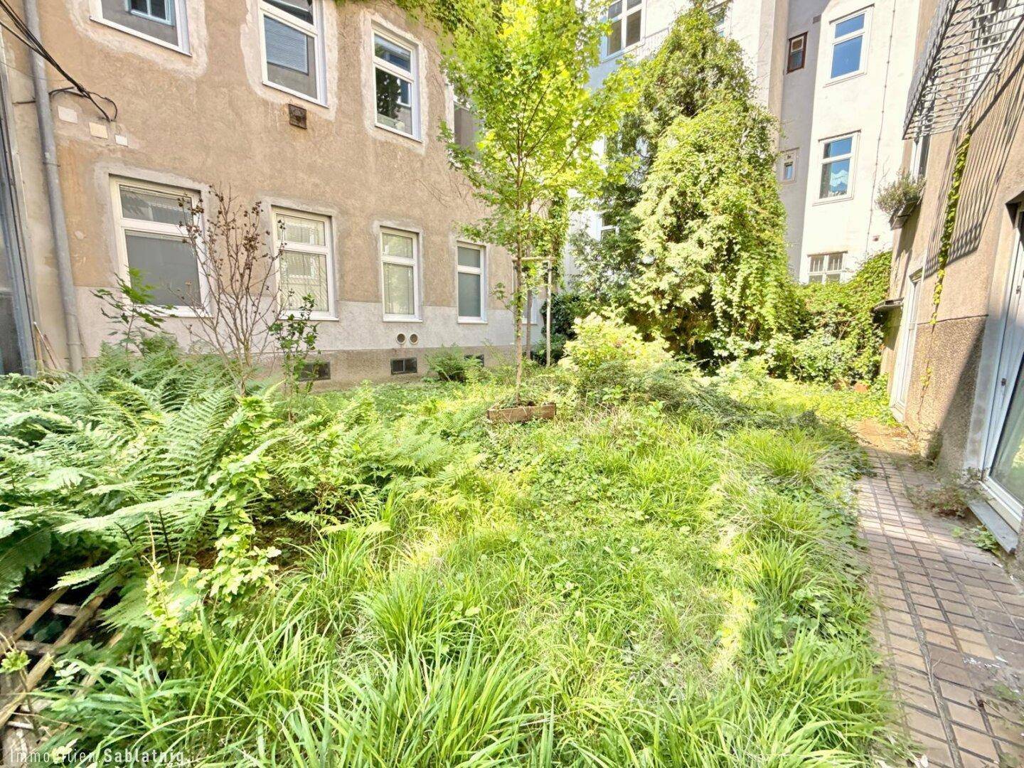Grüner Innenhofgarten mit üppiger Vegetation und einem gepflasterten Weg, umgeben von Gebäuden.