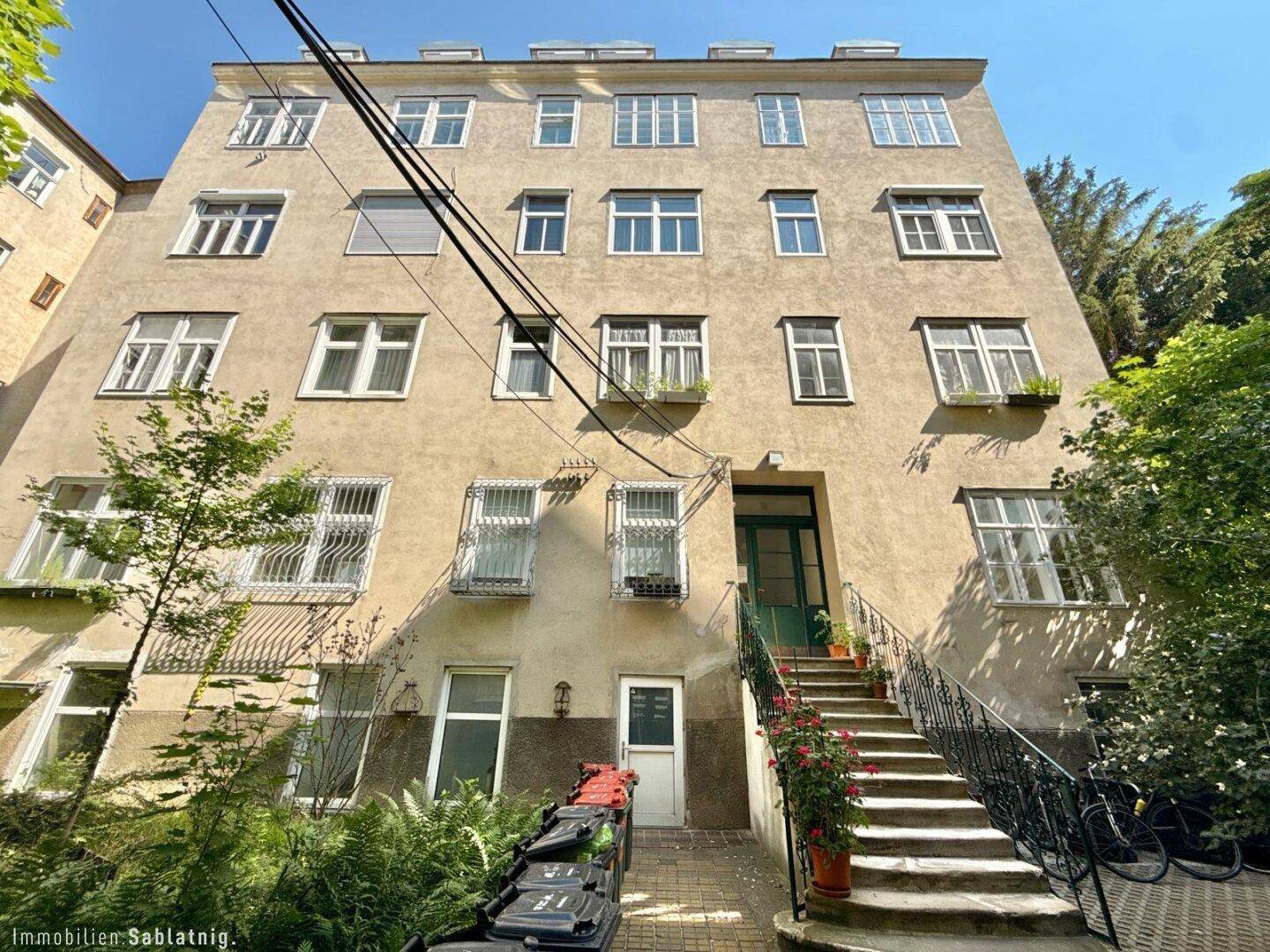Rückansicht eines Mehrfamilienhauses mit vielen Fenstern und einer Außentreppe, umgeben von Grün.