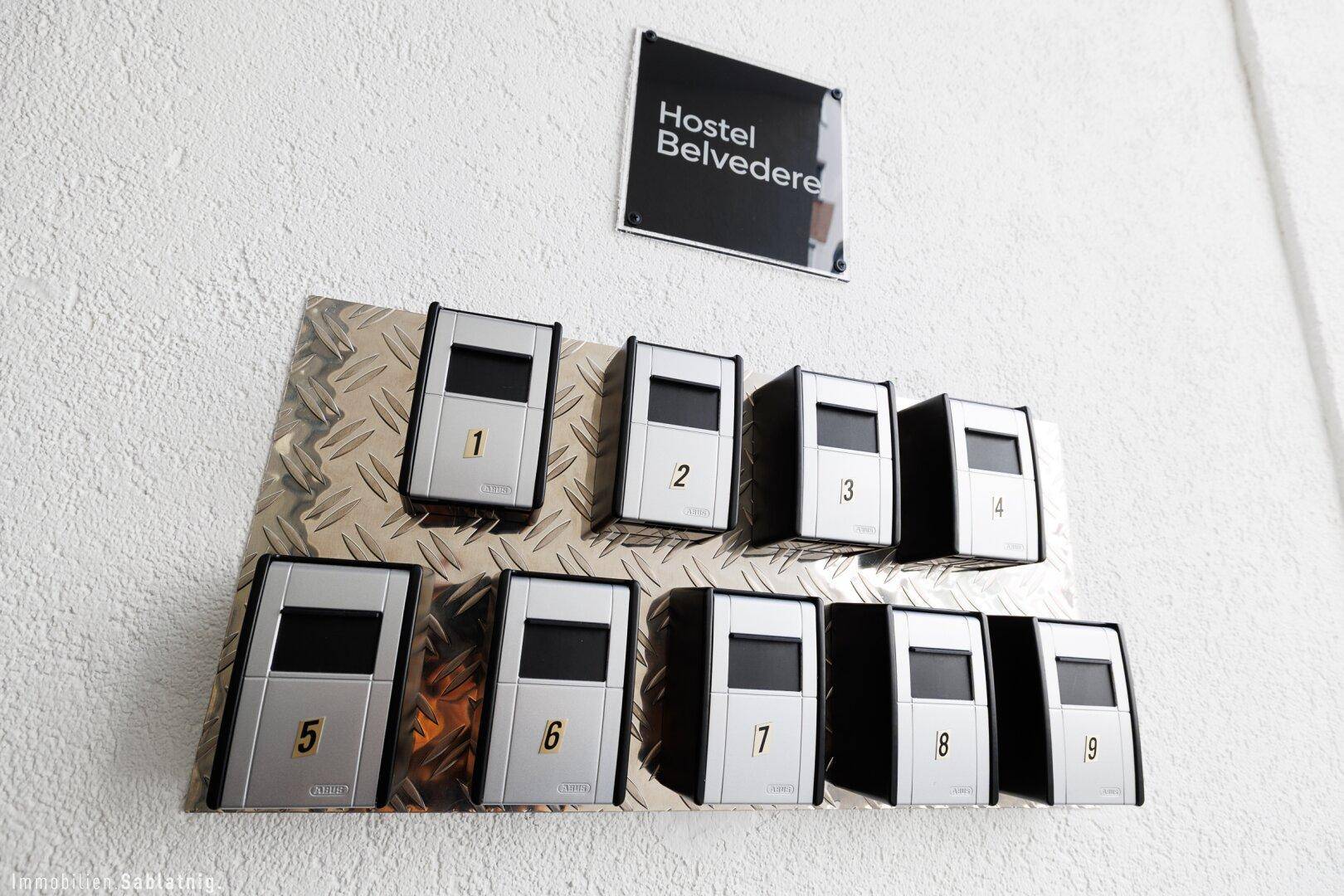 Reihe von neun Briefkästen oder Gegensprechanlagen an einer weißen Wand im Eingangsbereich des Hostels.