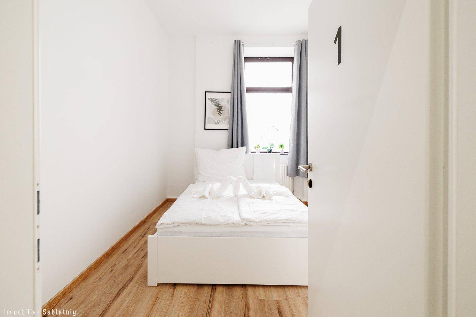 Helles Schlafzimmer mit Holzboden, weißem Bett und einem Fenster mit grauen Vorhängen für Privatsphäre.