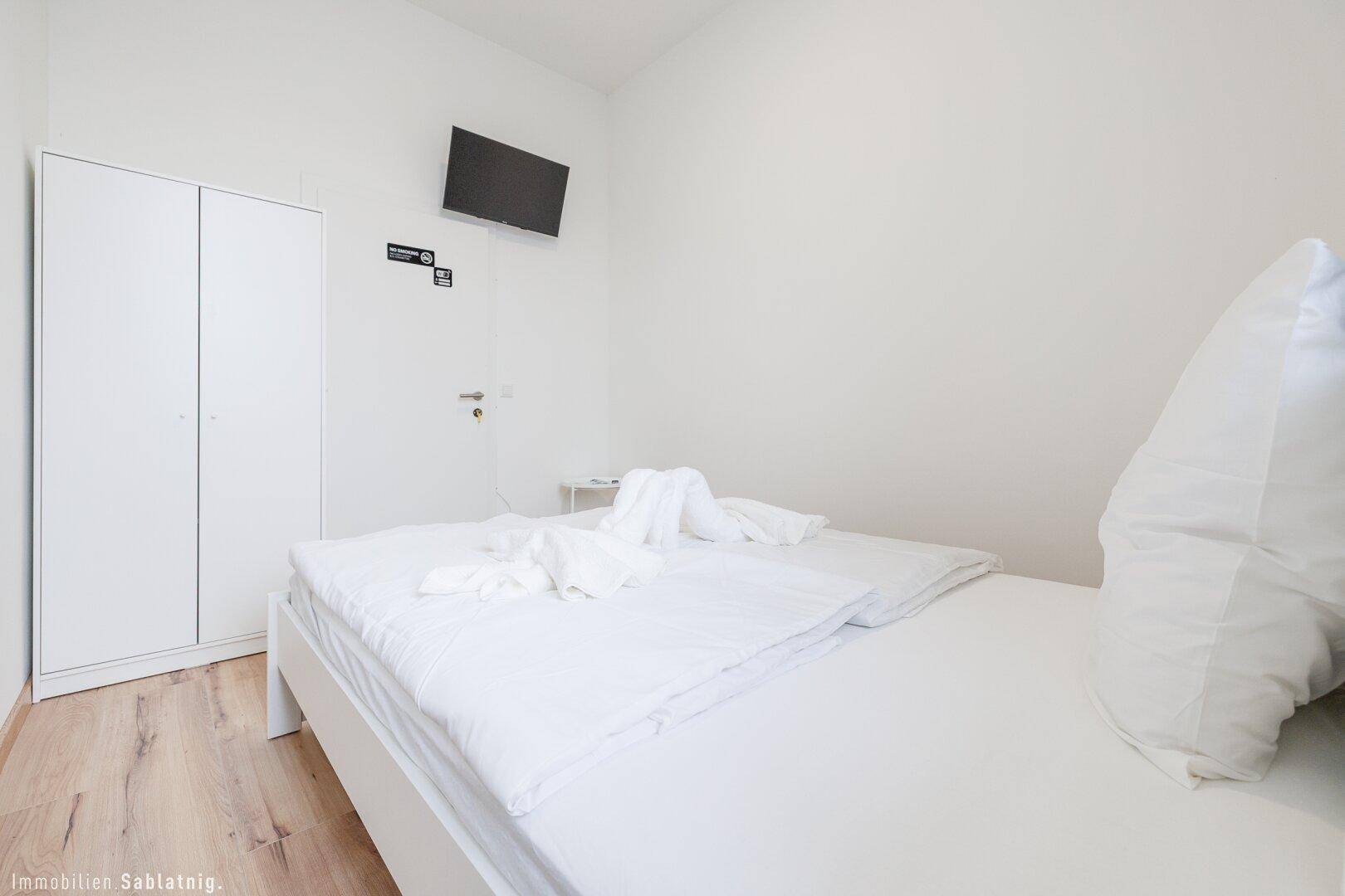 Schlafzimmer mit weißem Bett, Kleiderschrank und einem an der Wand montierten Flachbildfernseher.