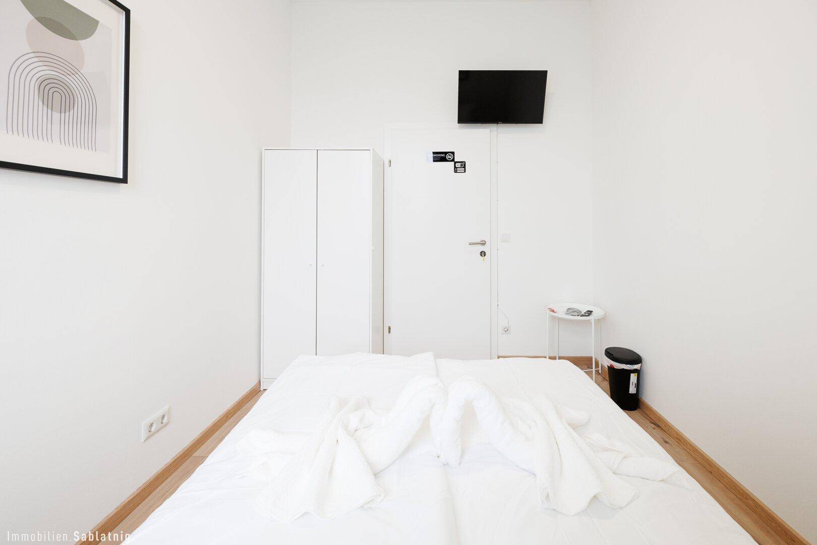 Minimalistisches Schlafzimmer mit weißem Bett, kleinem Beistelltisch und einem Fernseher an der Wand.