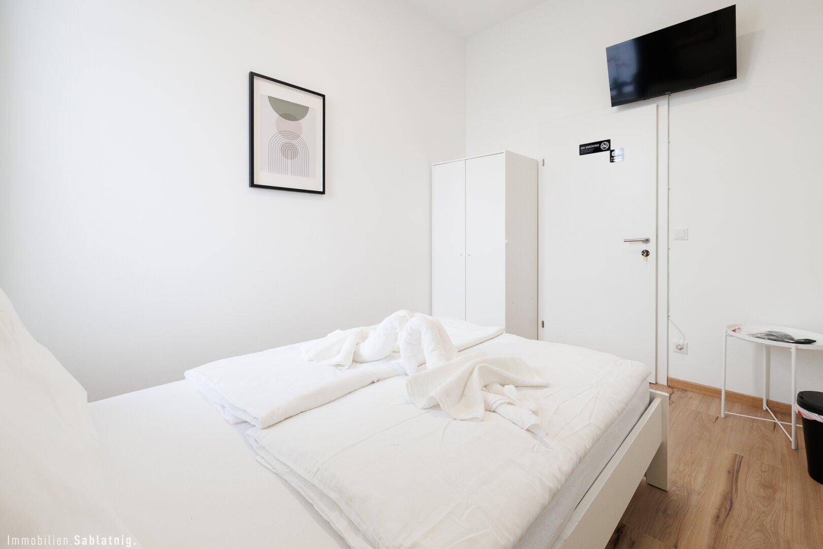 Helles Schlafzimmer mit weißem Bett, Holzboden und einem weißen Kleiderschrank für Stauraum.
