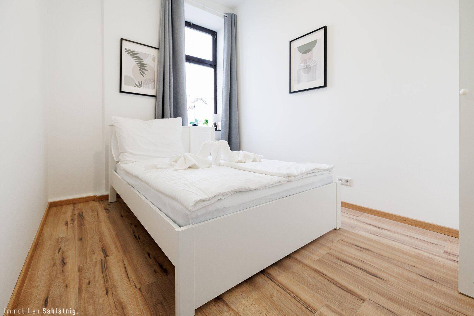 Helles Schlafzimmer mit Holzboden, weißem Bett und einem Fenster mit grauen Vorhängen für Privatsphäre.