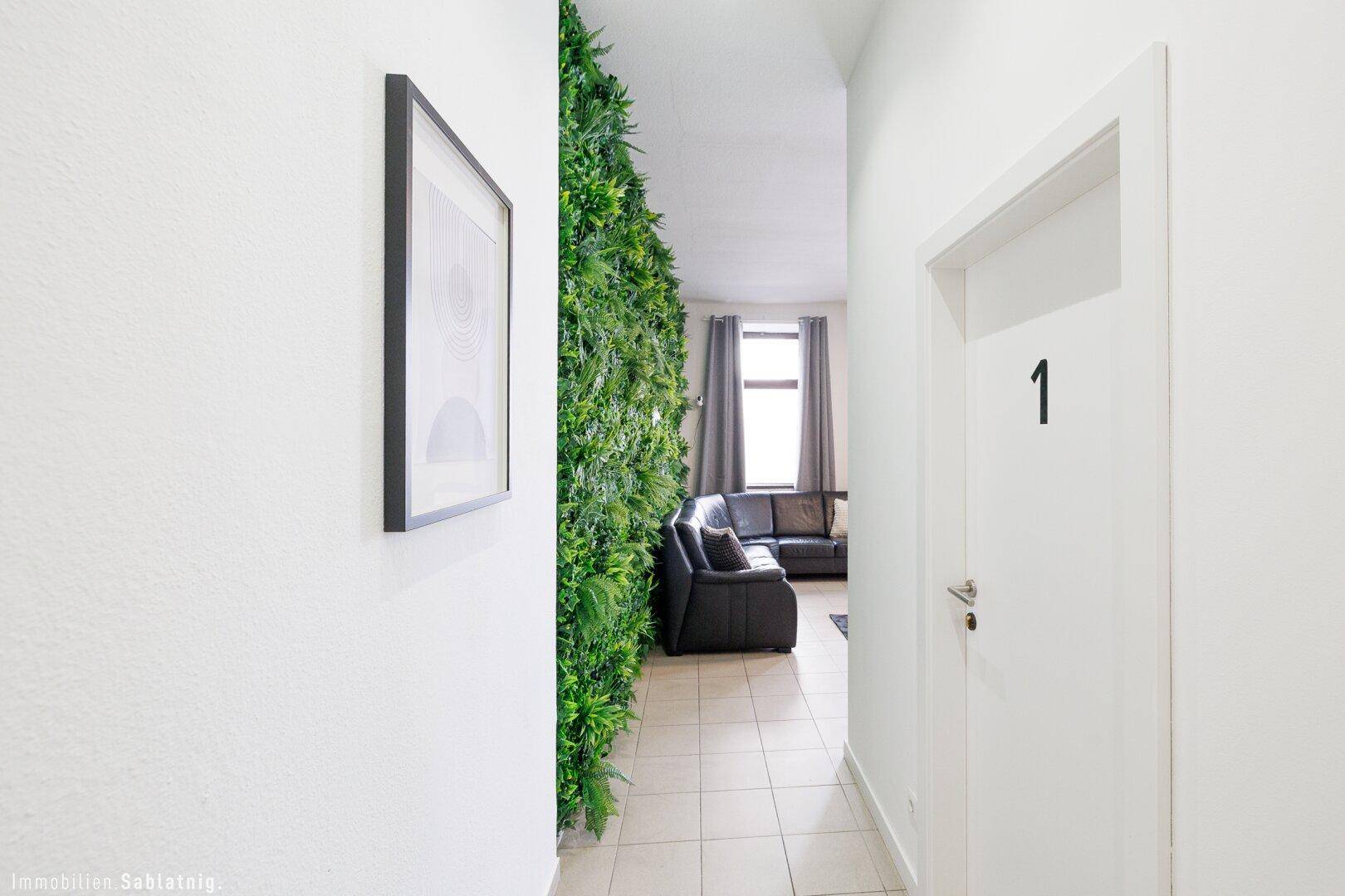 Heller Flur mit weißer Wand und einer grünen Pflanzenwand, die zum Gemeinschaftsbereich führt.