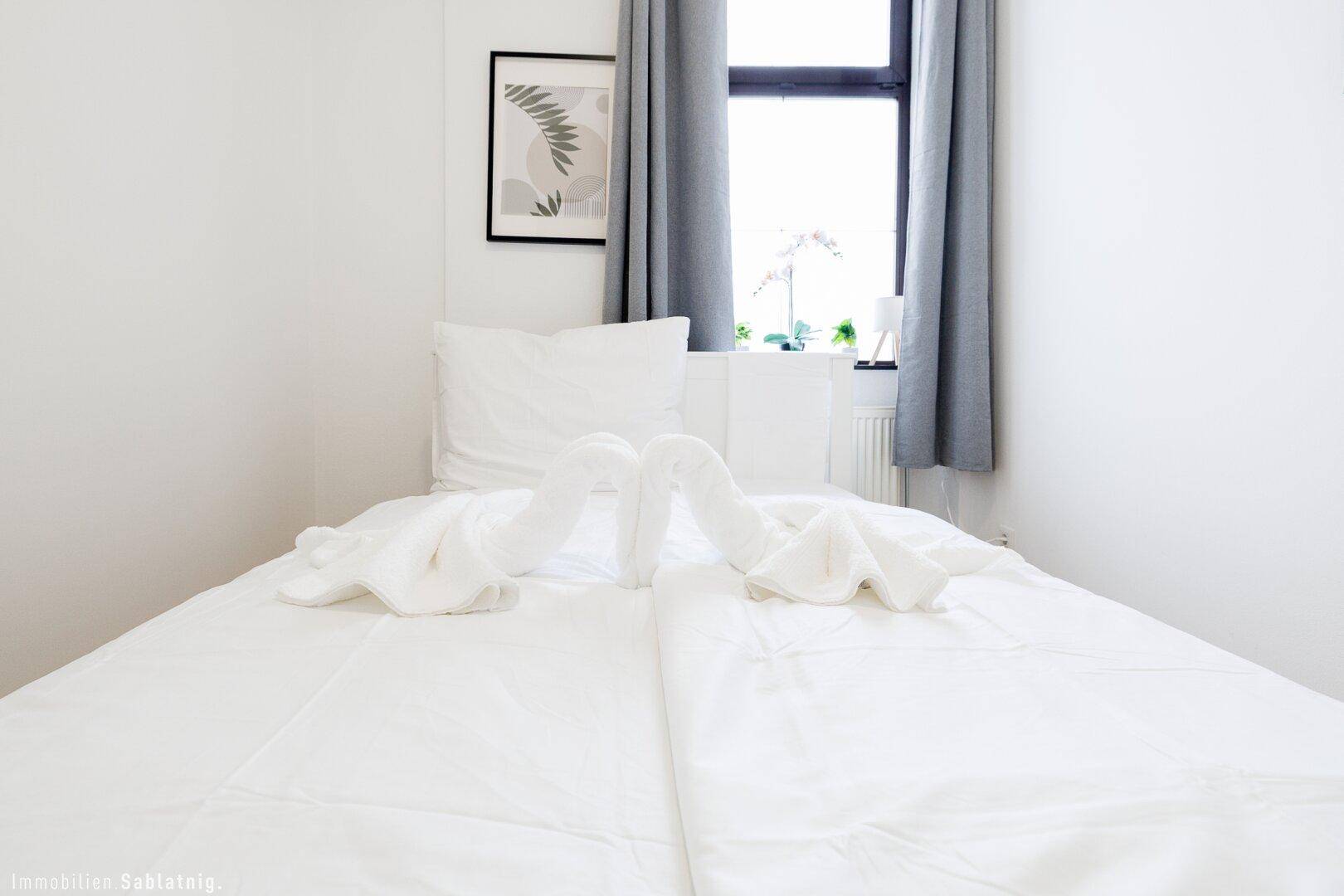 Helles Schlafzimmer mit Einzelbett, weißen Laken und grauen Vorhängen am Fenster.
