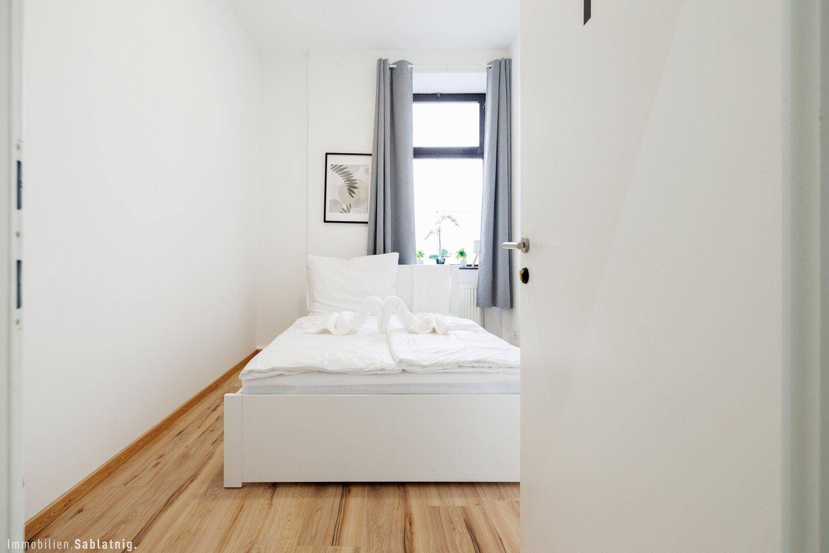 Blick in ein helles Schlafzimmer mit Einzelbett, Holzboden und einem Fenster mit Vorhängen.