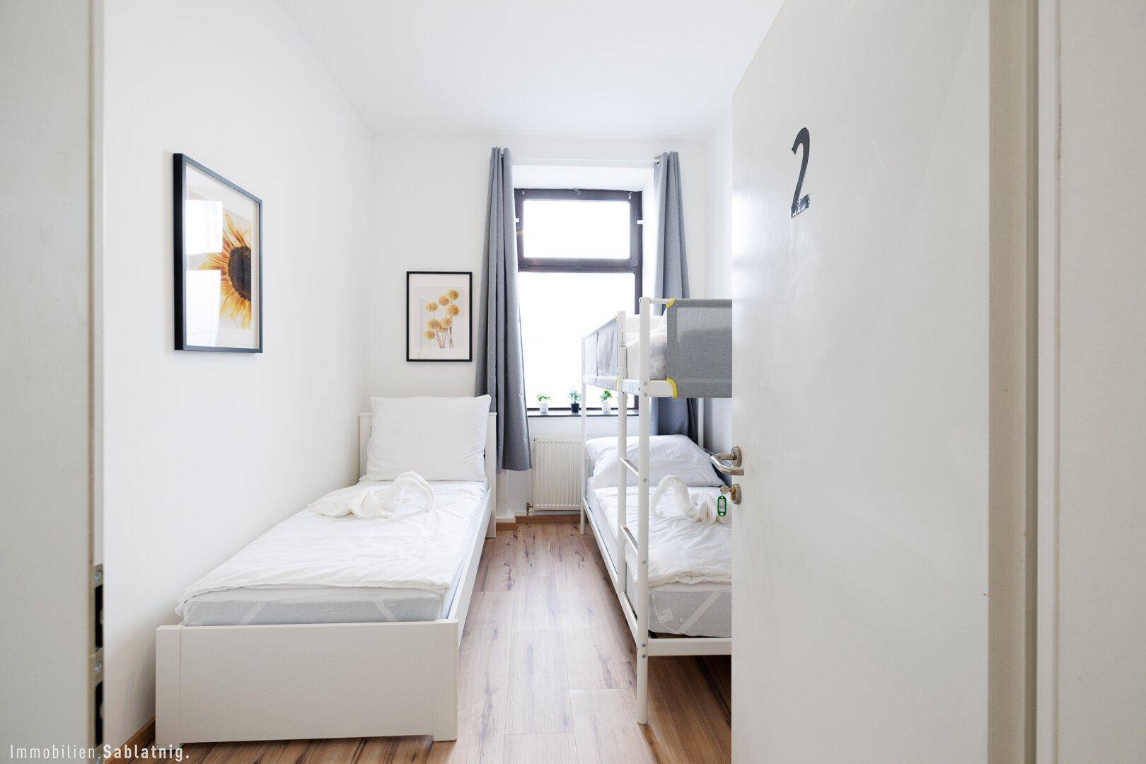 Schlafzimmer mit Einzelbett und Etagenbett, Holzboden und einem Fenster mit grauen Vorhängen.