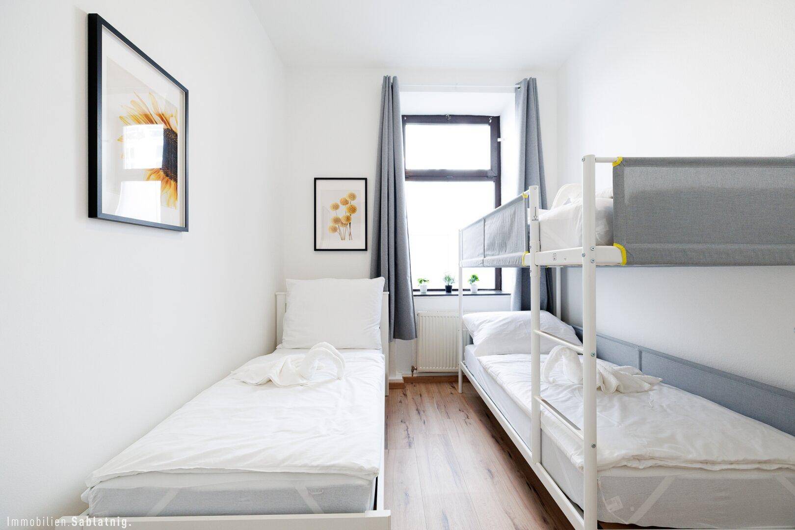 Helles Schlafzimmer mit Einzelbett und Etagenbett, gerahmten Bildern und einem Fenster.