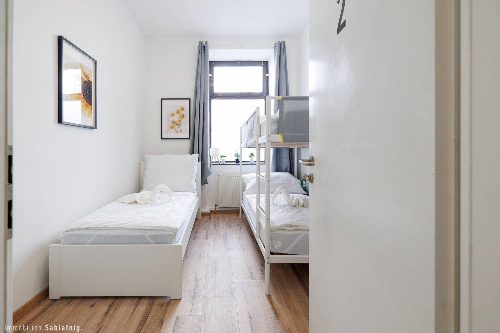 Blick in ein Schlafzimmer mit Einzelbett und Etagenbett, Holzboden und einem Fenster.