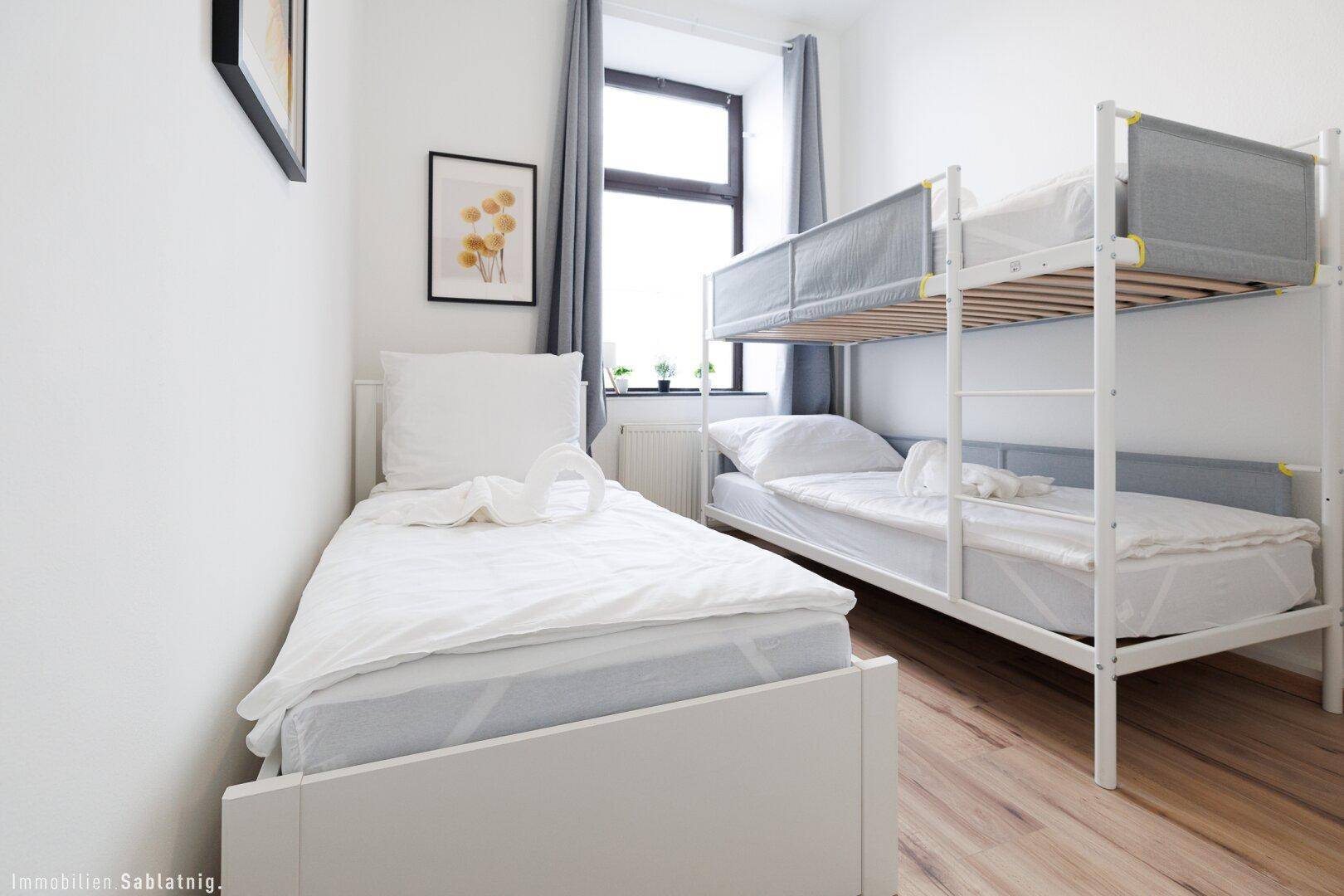 Schlafzimmer mit Einzelbett und Etagenbett, weißen Wänden und einem Fenster mit Vorhängen.