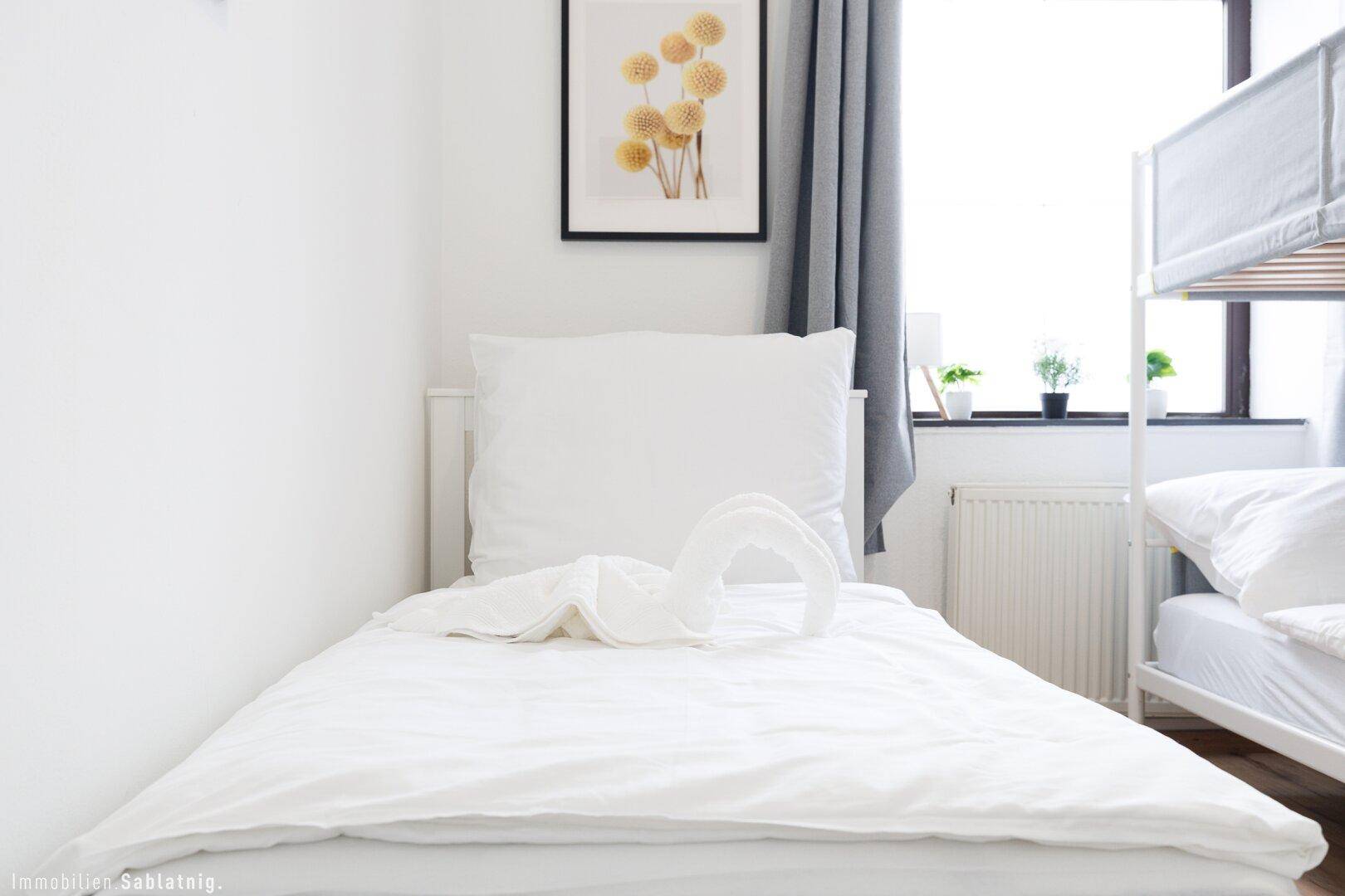 Nahaufnahme eines Einzelbettes in einem Schlafzimmer mit Etagenbett und Fenster im Hintergrund.