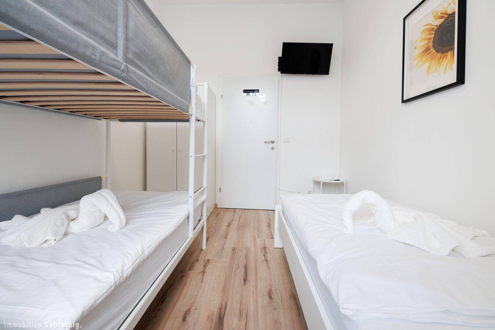 Schlafzimmer mit Etagenbett und Einzelbett, Wandfernseher und weißem Kleiderschrank.