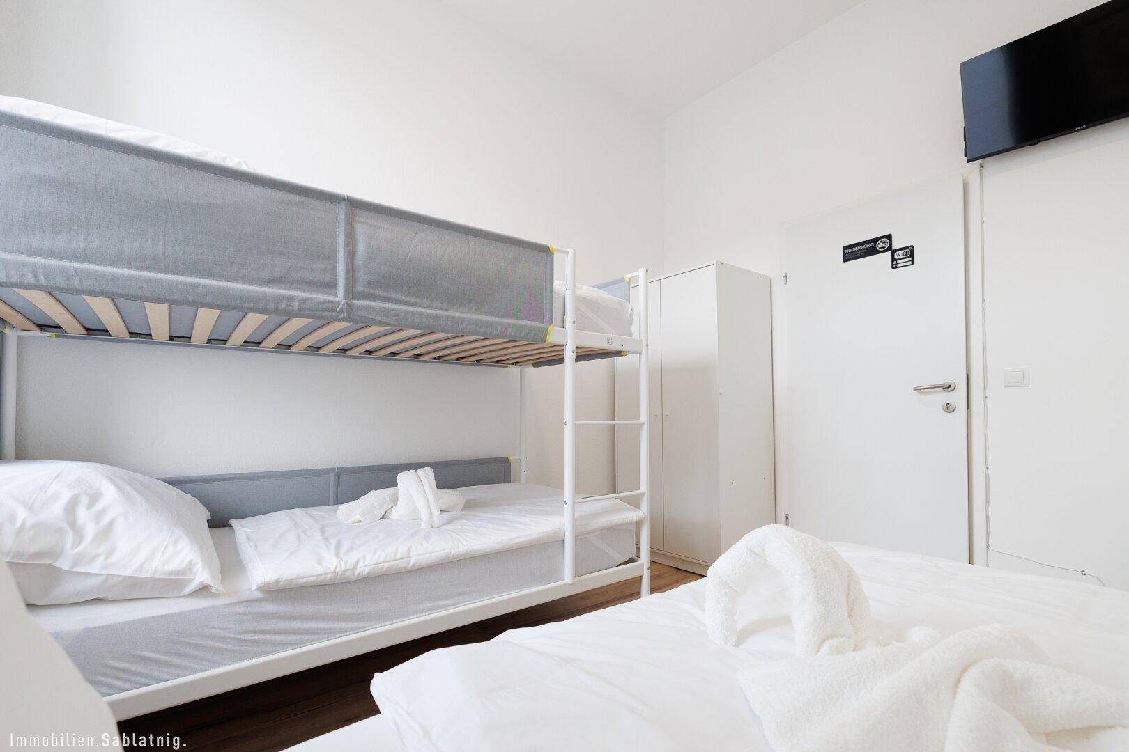 Schlafzimmer mit Etagenbett und Einzelbett, Wandfernseher und weißem Kleiderschrank.