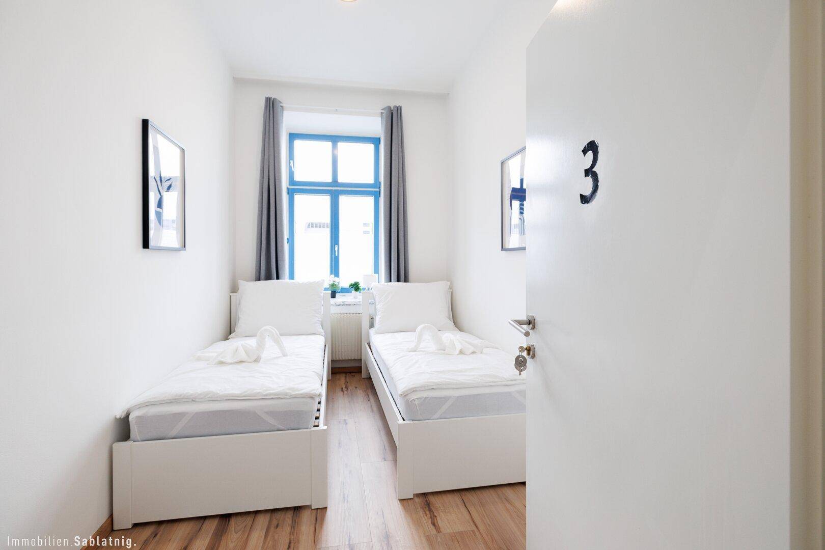 Helles Schlafzimmer mit zwei Einzelbetten, Holzboden und einem Fenster mit blauen Rahmen.