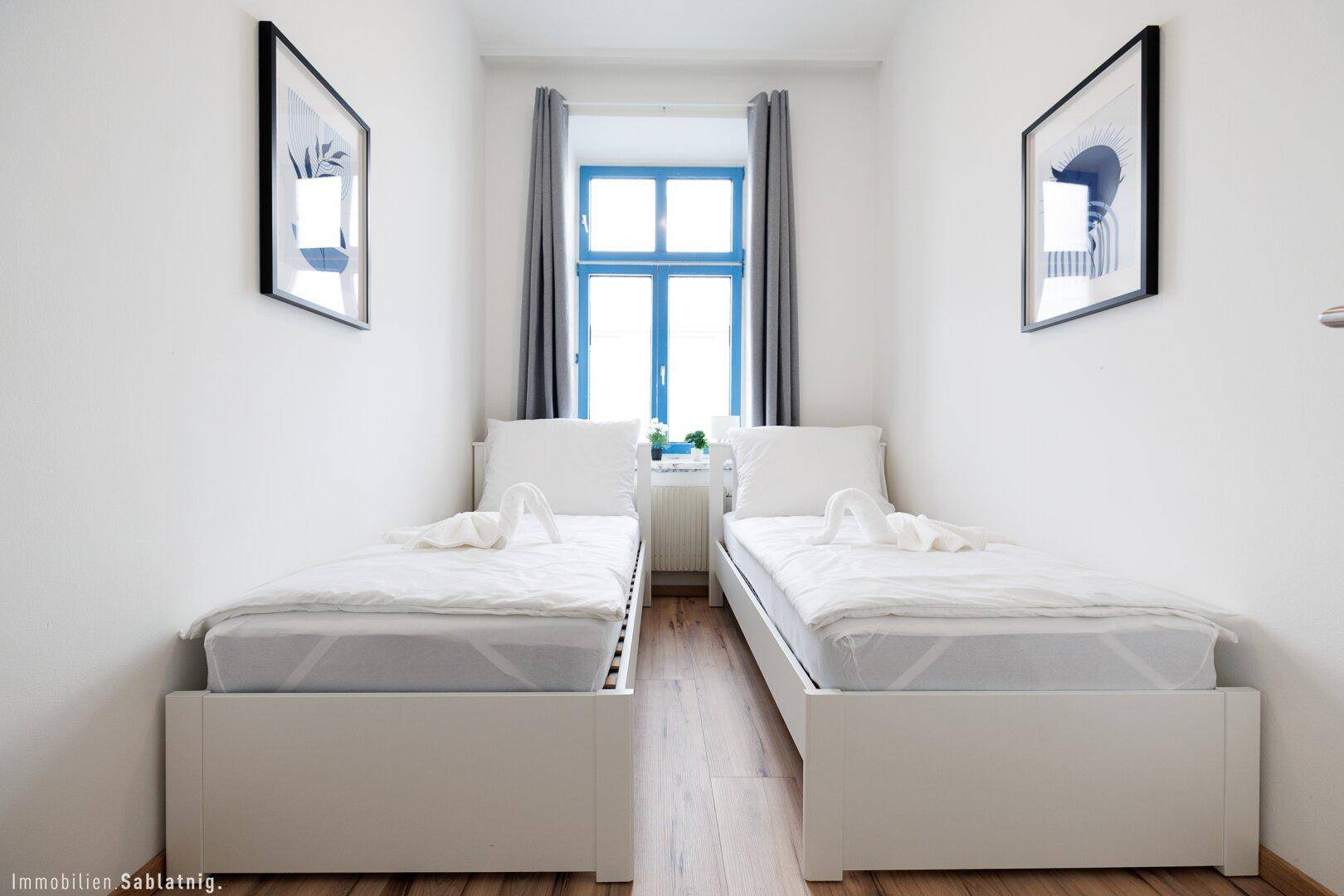 Schlafzimmer mit zwei Einzelbetten, weißen Wänden und einem Fenster mit blauen Rahmen.