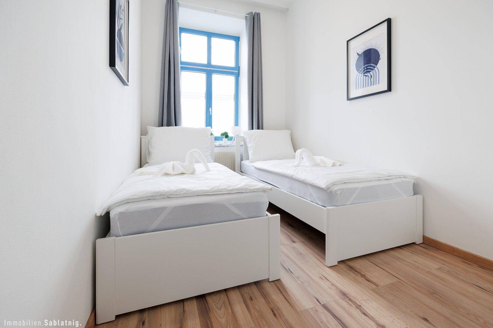 Schlafzimmer mit zwei Einzelbetten, Holzboden und einem Fenster mit blauen Rahmen.