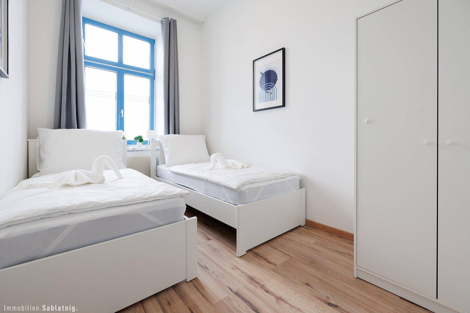 Schlafzimmer mit zwei Einzelbetten, weißem Kleiderschrank und einem Fenster mit blauen Rahmen.