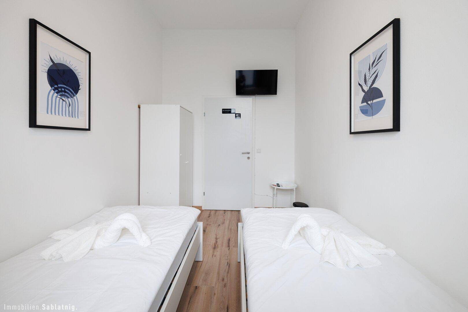 Schlafzimmer mit zwei Einzelbetten, Wandfernseher und weißem Kleiderschrank.