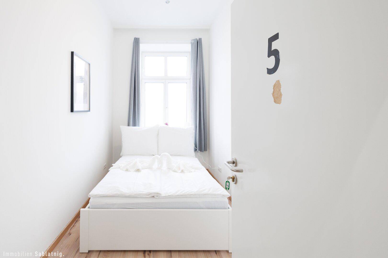 Blick in ein helles Schlafzimmer mit Einzelbett, Holzboden und einem Fenster mit Vorhängen.