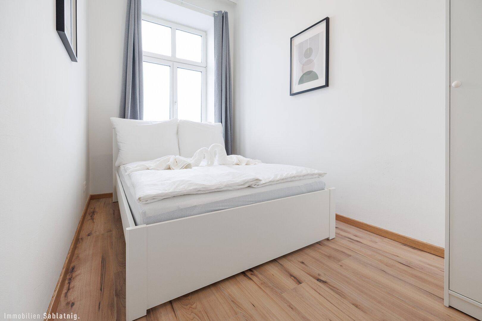 Helles Schlafzimmer mit einem Doppelbett, Holzboden und einem Fenster mit grauen Vorhängen.
