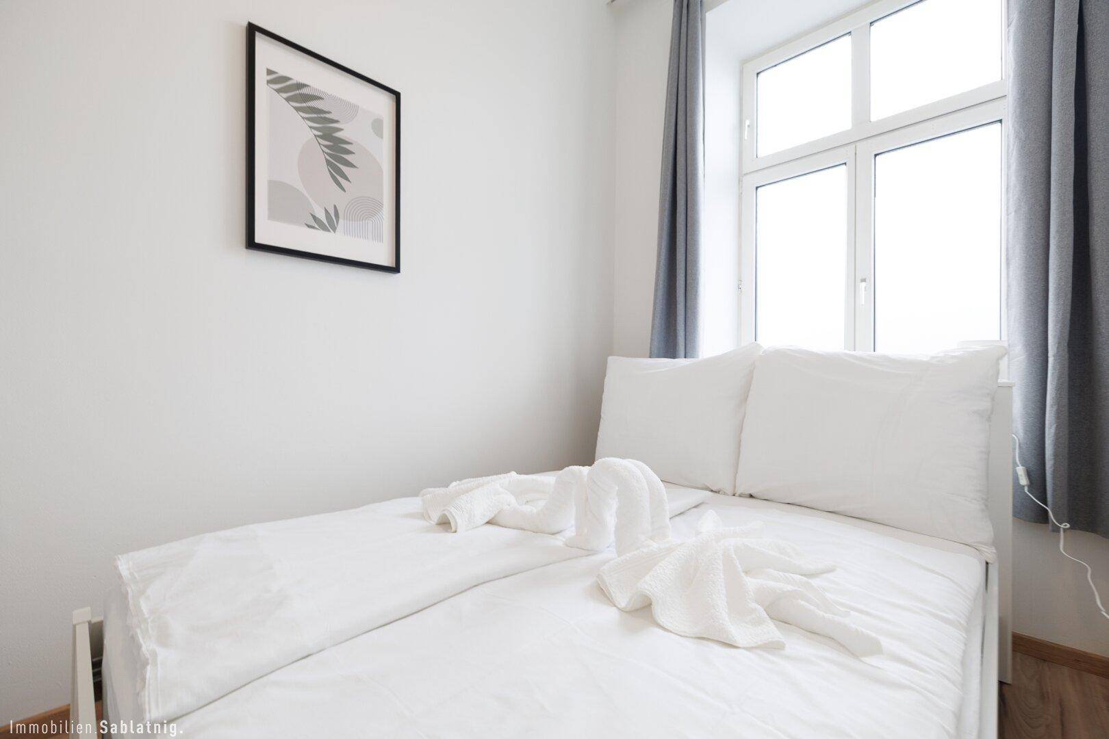 Modern eingerichtetes Schlafzimmer mit weißem Bett, Holzboden und einem Fenster mit grauen Vorhängen.