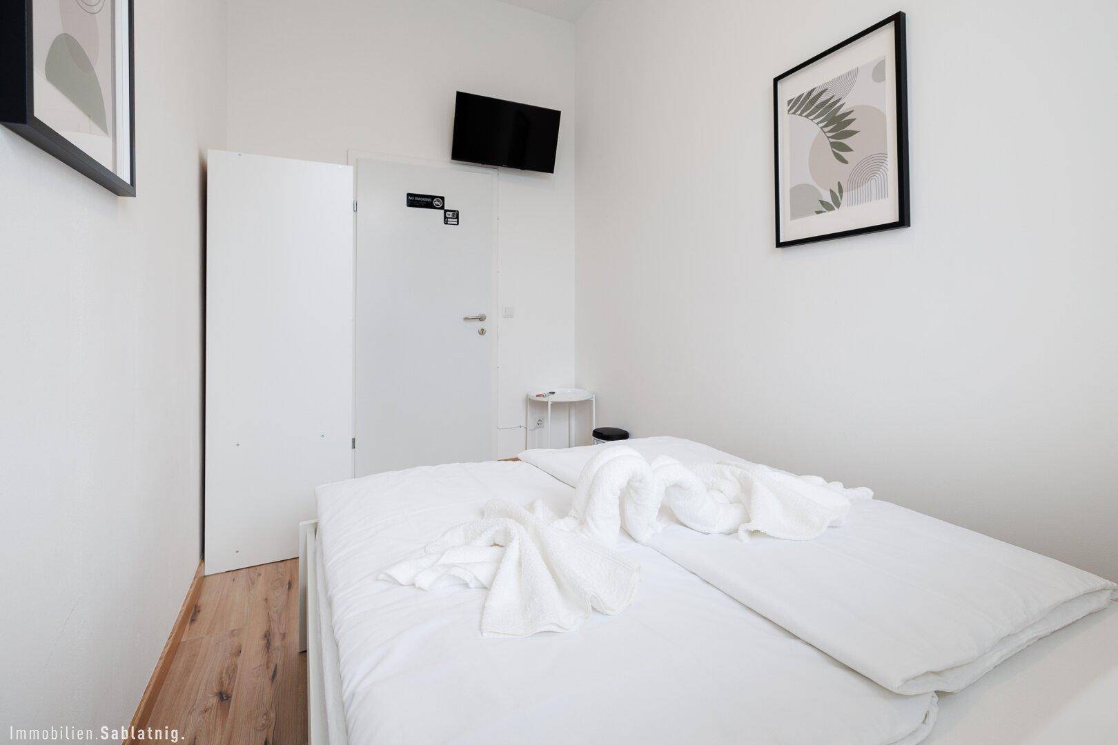 Schlafzimmer mit Doppelbett, Holzboden, einem an der Wand montierten Fernseher und einer weißen Tür.