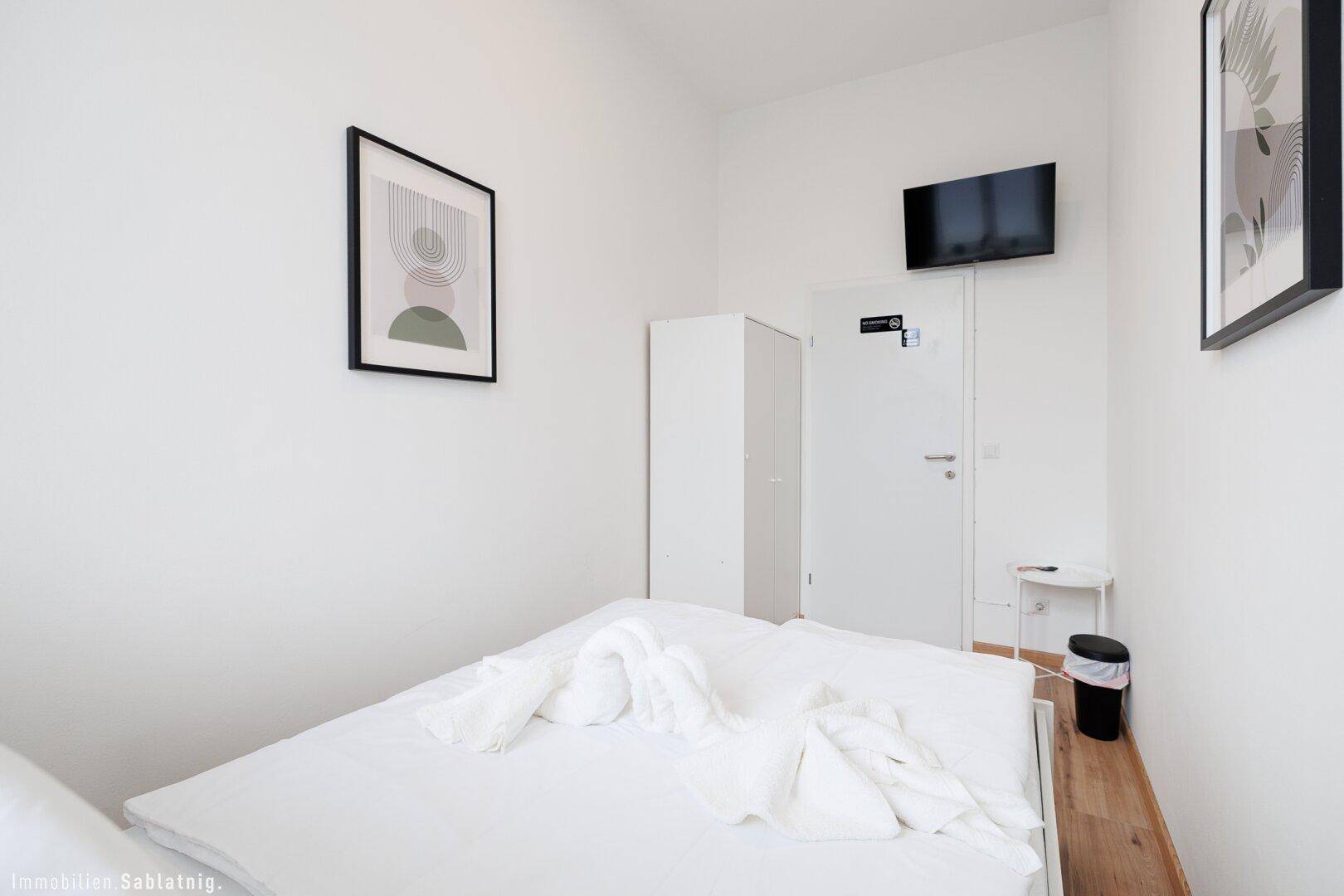 Schlafzimmer mit Doppelbett, Holzboden, einem an der Wand montierten Fernseher und einem kleinen Beistelltisch.