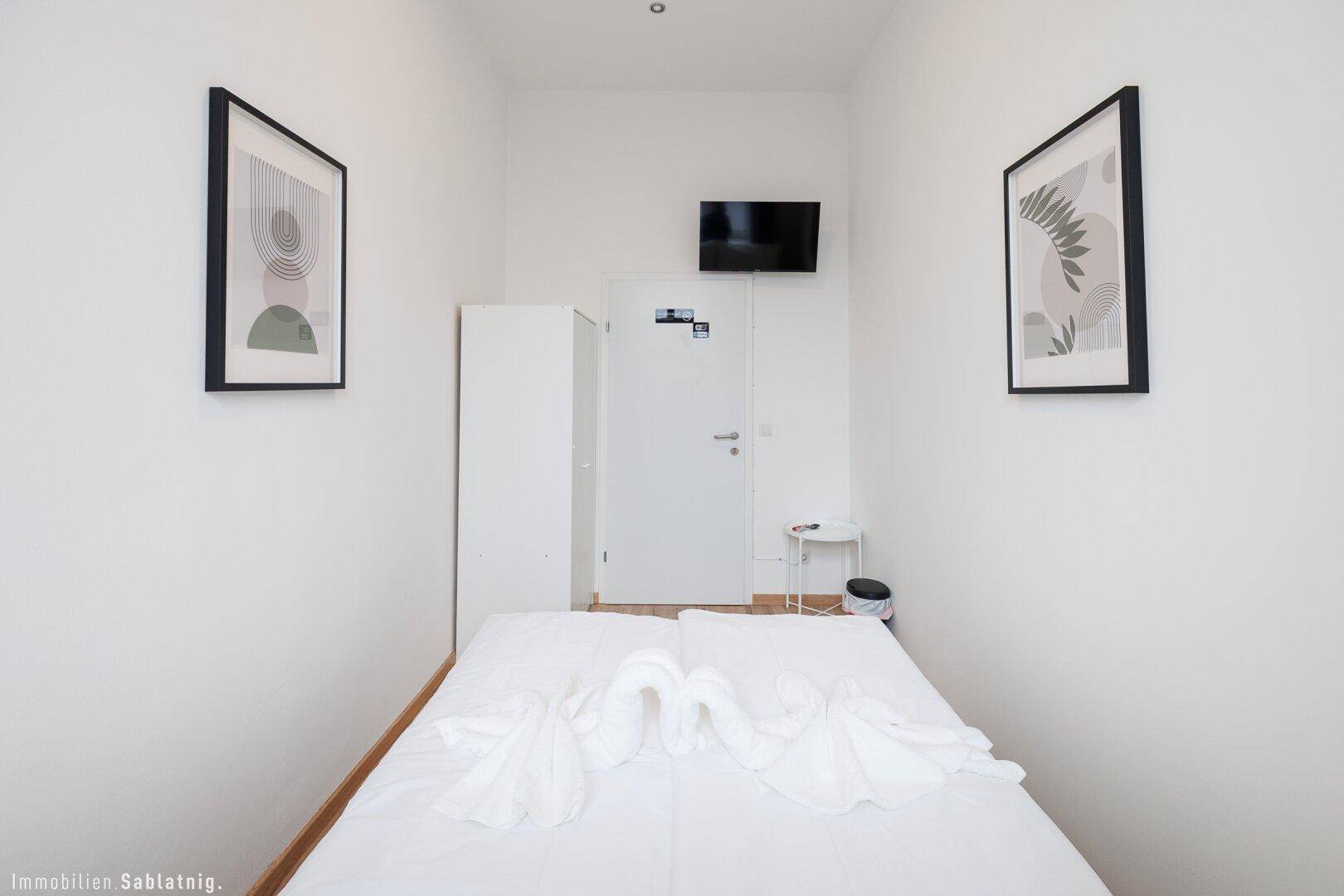 Helles Schlafzimmer mit Doppelbett, Holzboden und einem an der Wand montierten Fernseher.