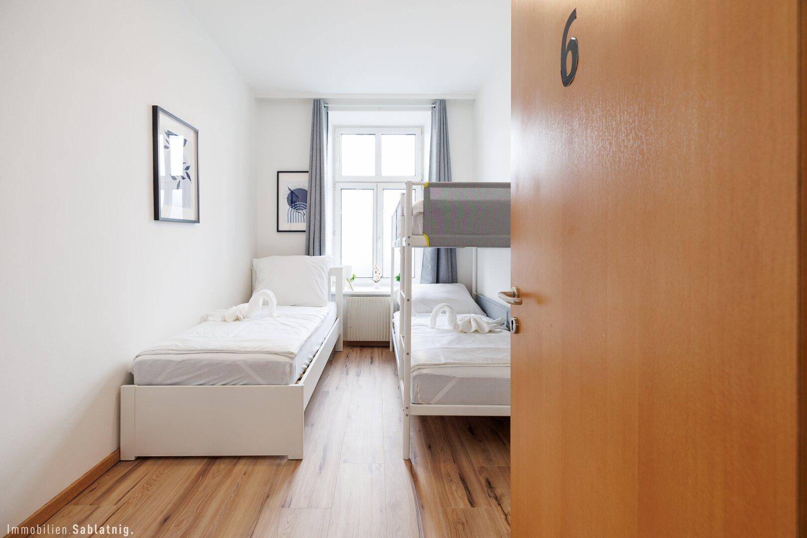 Schlafzimmer mit Einzelbett und Etagenbett, Holzboden und einem Fenster mit grauen Vorhängen.