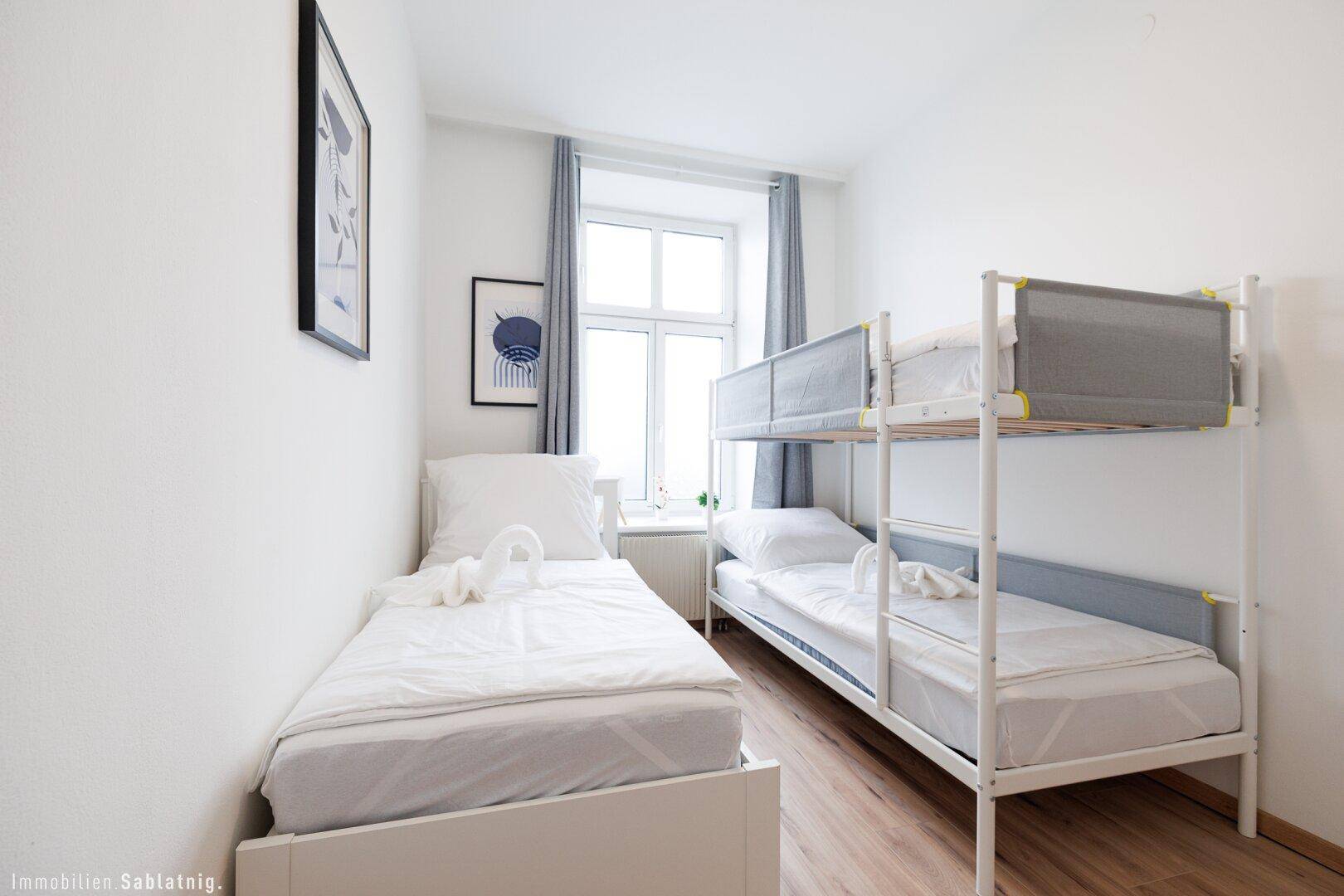 Helles Schlafzimmer mit Einzelbett und Etagenbett, Holzboden und einem Fenster.