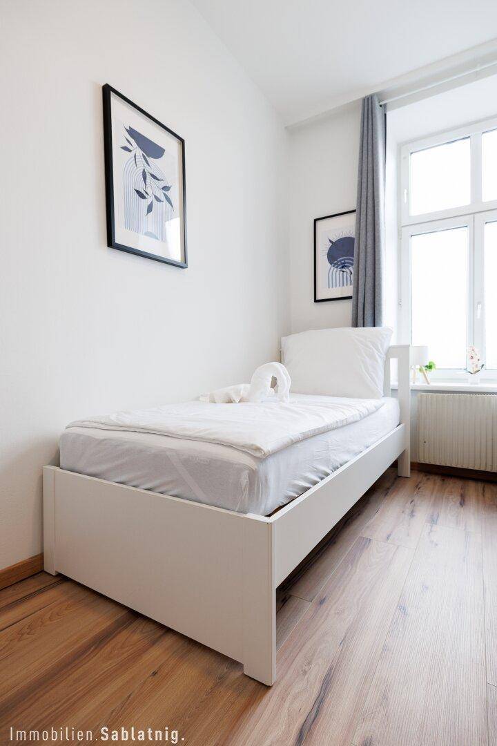 Einzelbett in einem hellen Schlafzimmer mit Holzboden, Fenster und grauen Vorhängen.