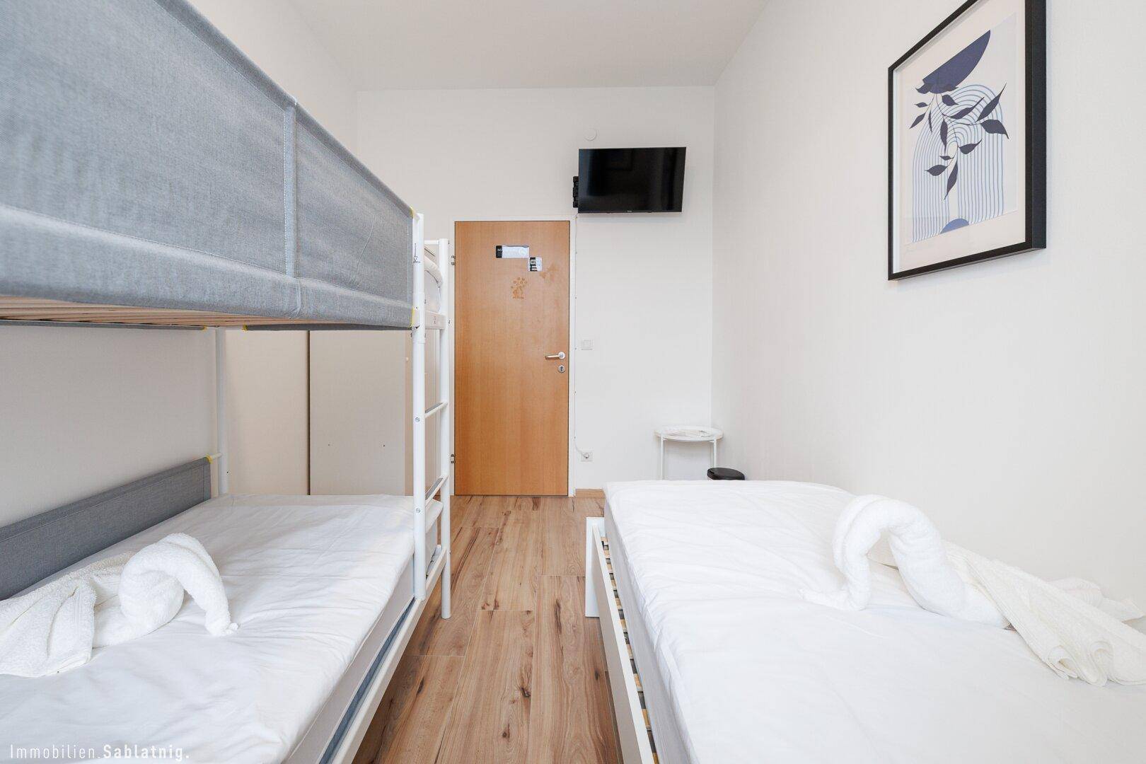 Schlafzimmer mit Einzelbett und Etagenbett, Holzboden und einem an der Wand montierten Fernseher.