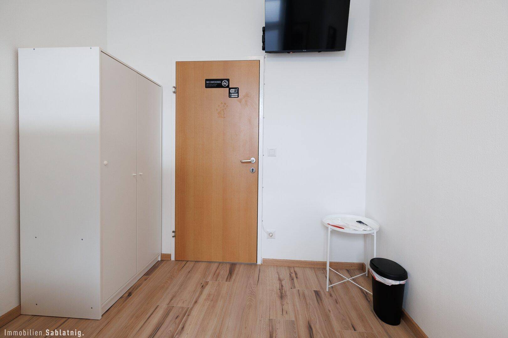 Bereich neben dem Bett mit einem Kleiderschrank, einer Holztür und einem an der Wand montierten Fernseher.