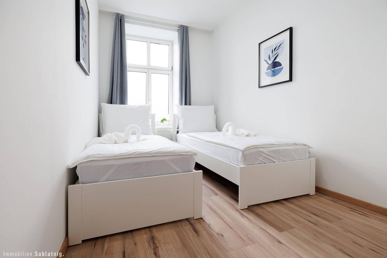 Schlafzimmer mit zwei Einzelbetten, Holzboden und einem Fenster, das viel Tageslicht hereinlässt.