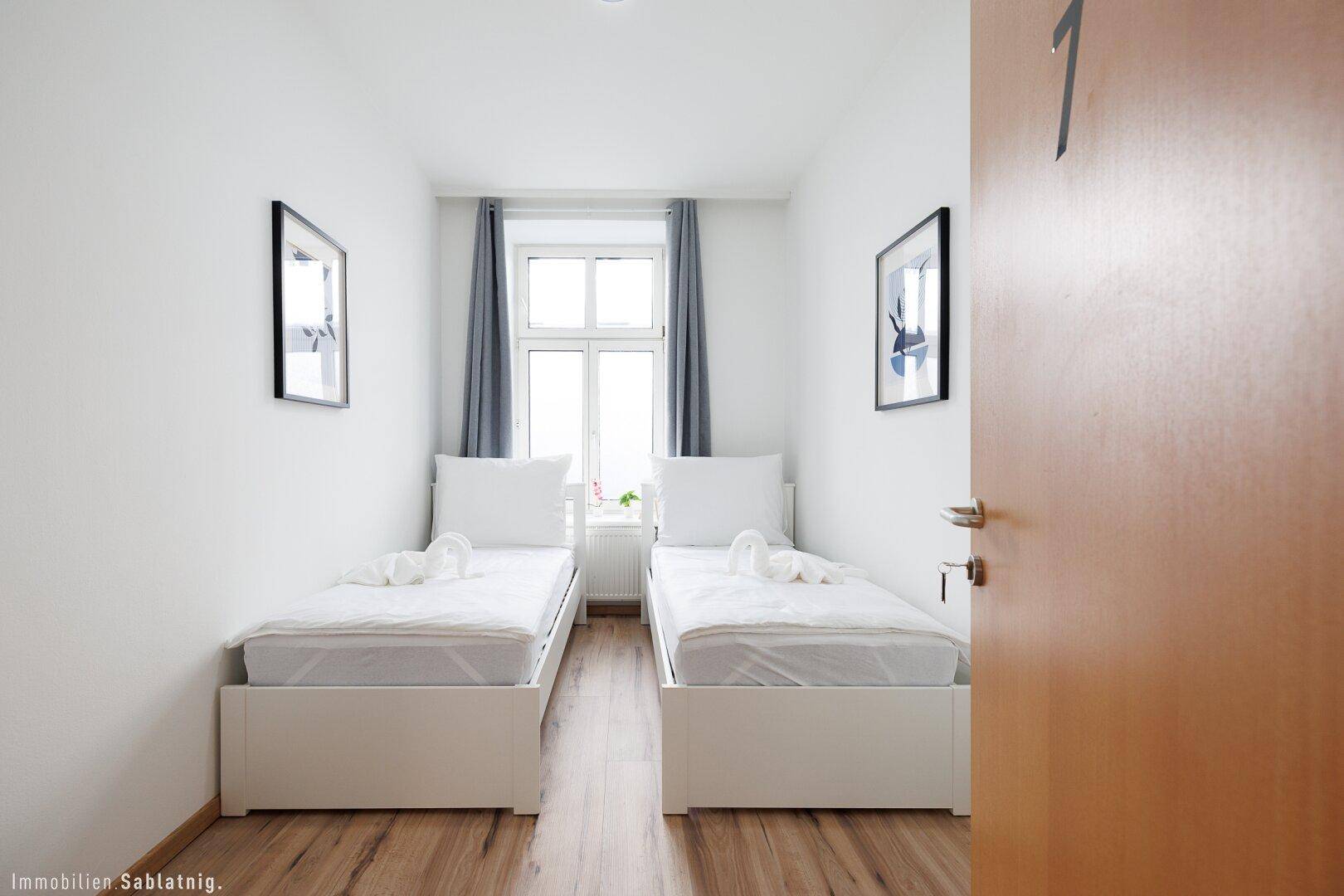 Helles Schlafzimmer mit zwei Einzelbetten, Holzboden und einem Fenster mit grauen Vorhängen.
